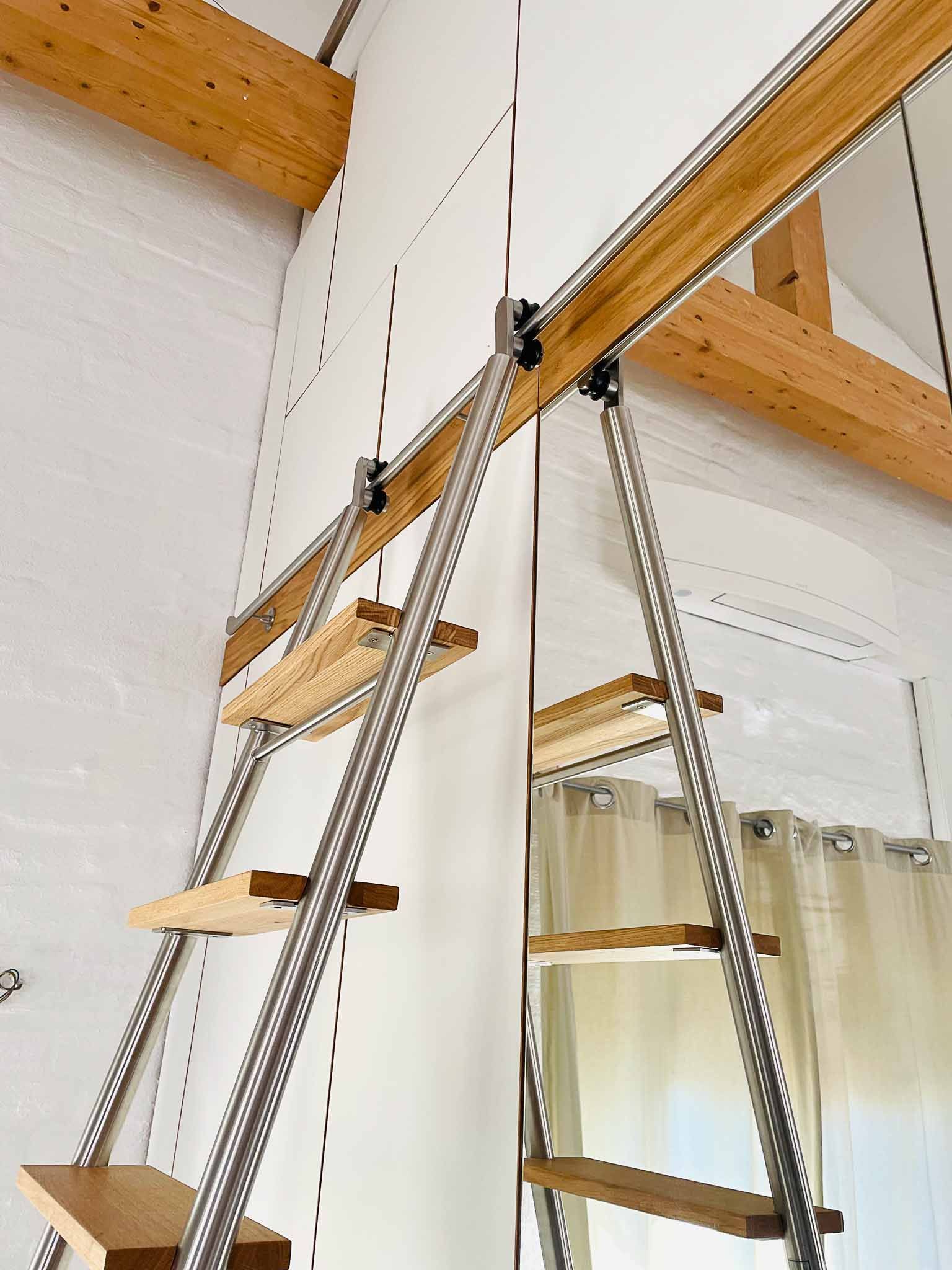 An einer Wand mit Metallschienen befestigte Holzleiter; natürliche Holztöne, cremefarbene Wand, im Hintergrund Vorhang.