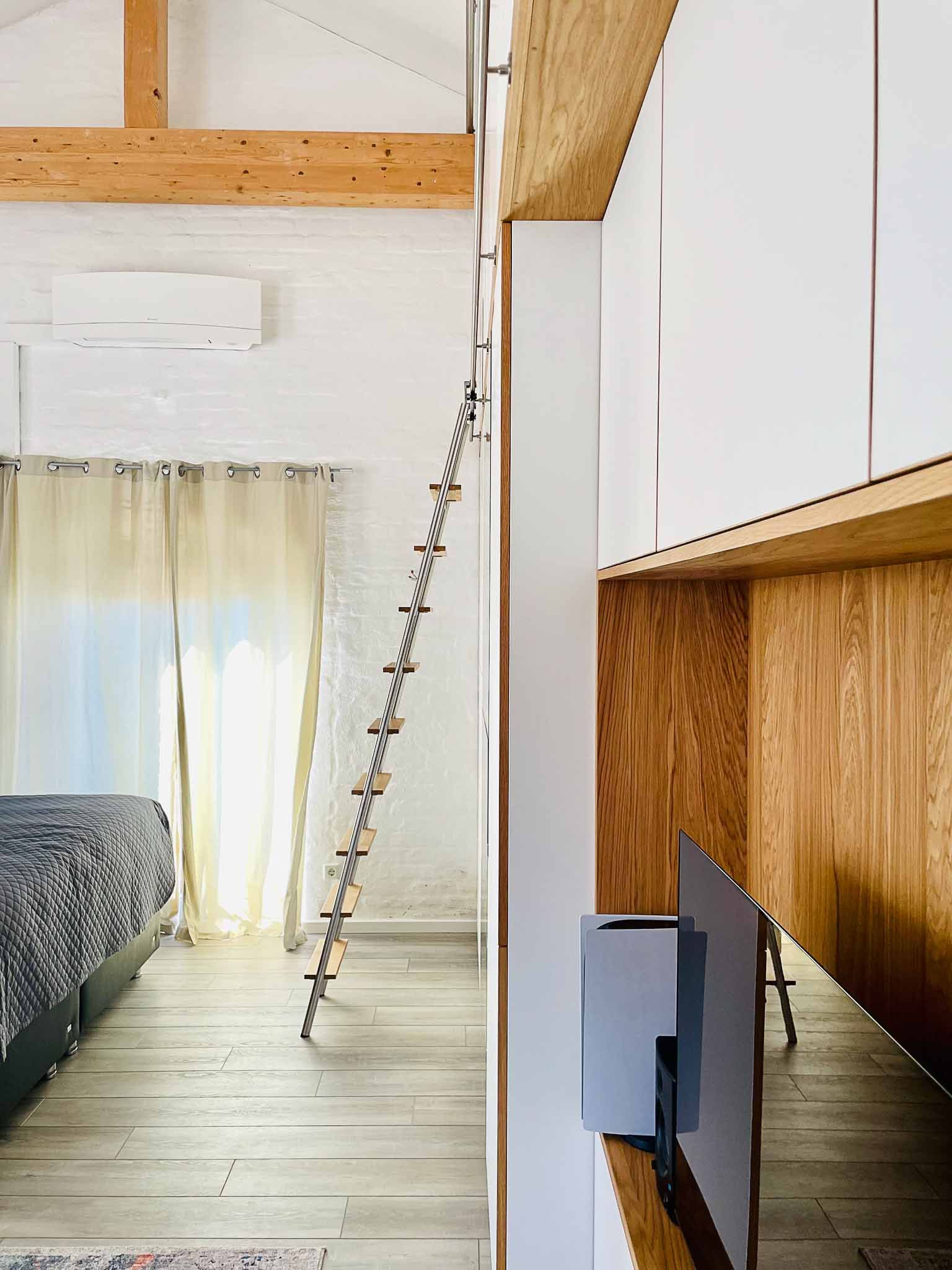 Schlafzimmereinrichtung mit einer Leiter, die zu einem Dachboden führt, weißen Ziegelwänden, Holzbalken und einem Fenster mit Vorhängen.