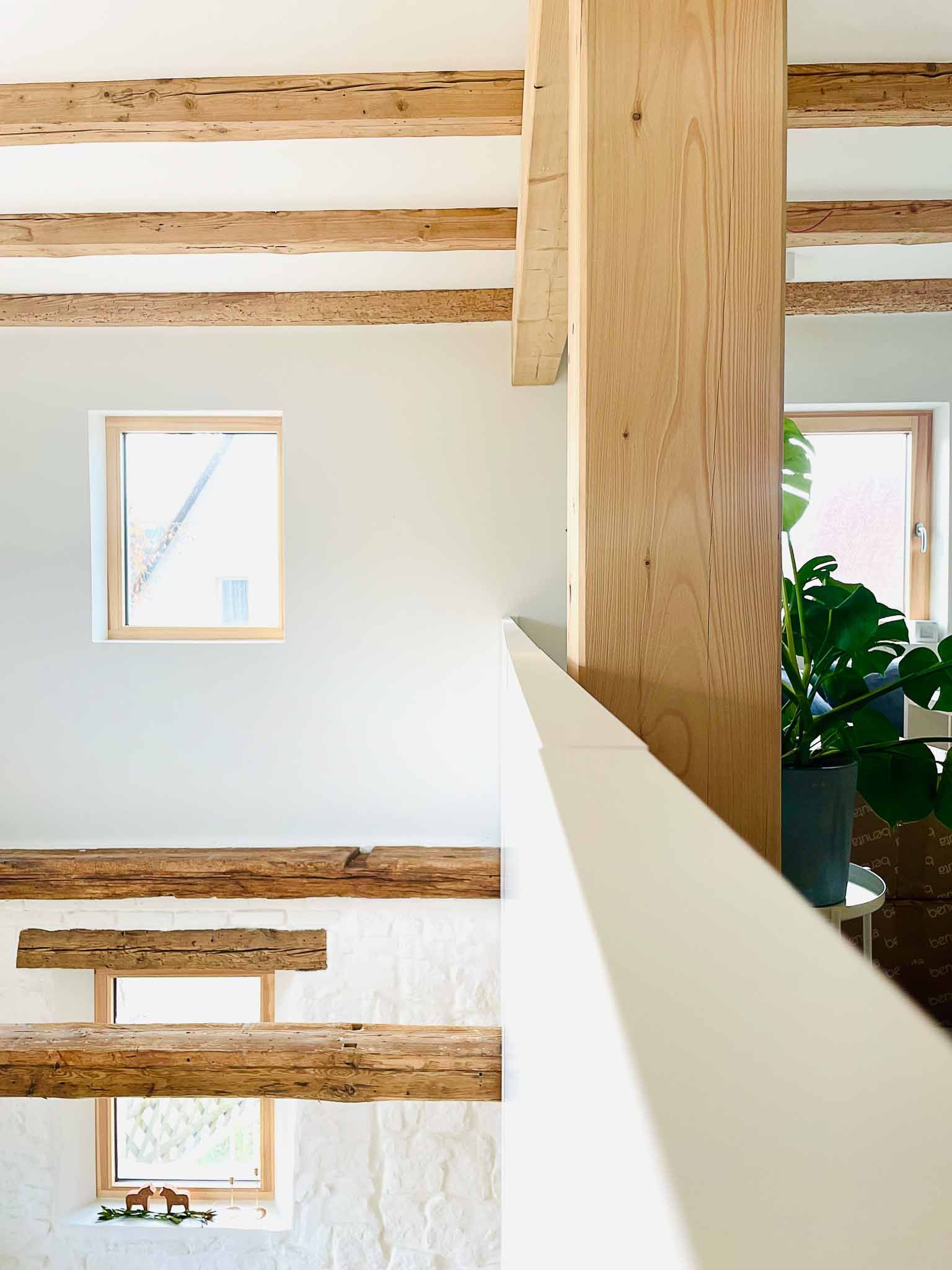 Innenansicht eines hellen Raums mit freiliegenden Holzbalken und einem weißen Geländer sowie einem Fenster.