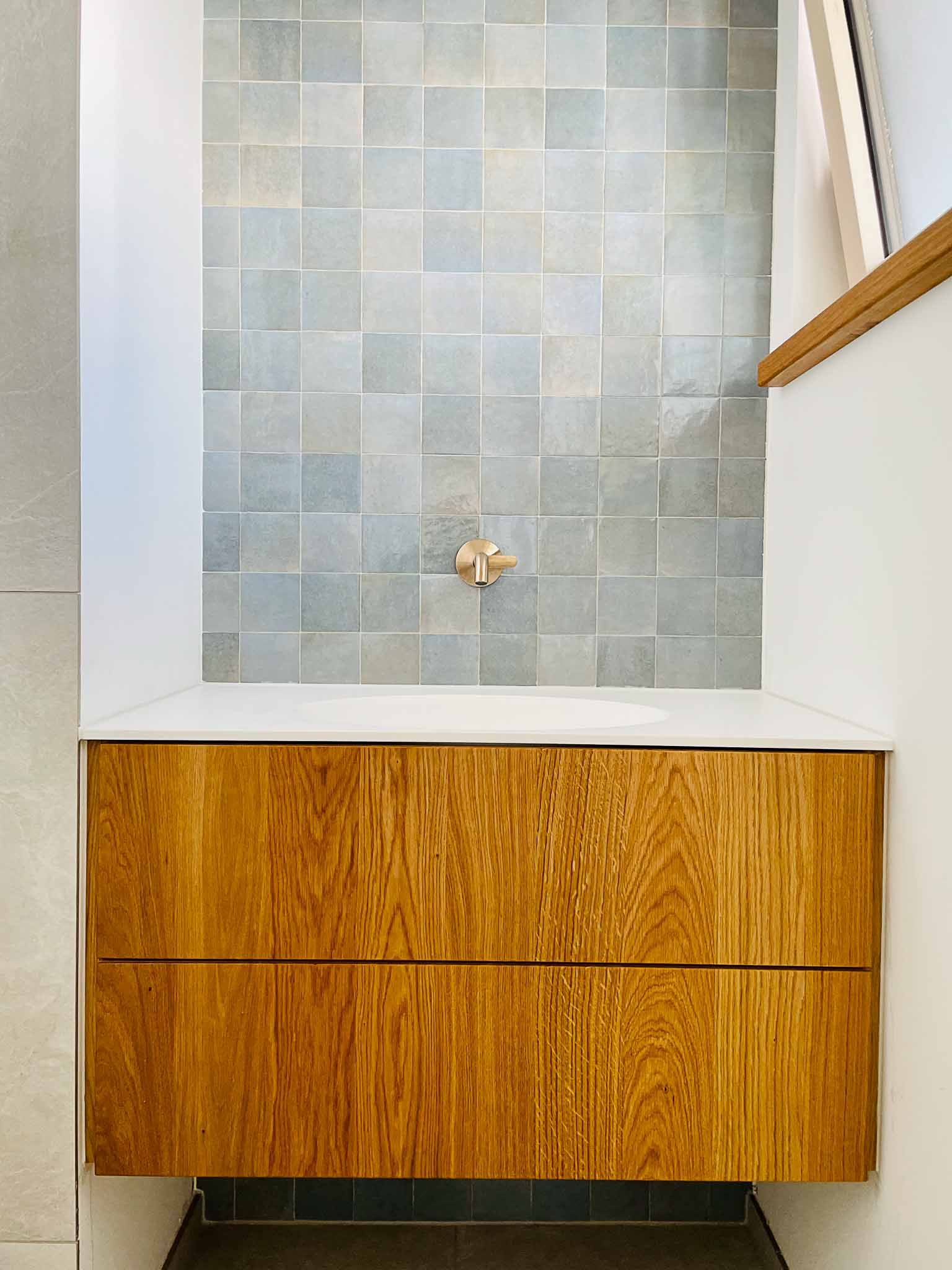 Badezimmerwaschtisch aus Holz mit weißer Arbeitsplatte und einer blaugrauen Duschwand mit quadratischen Fliesen.