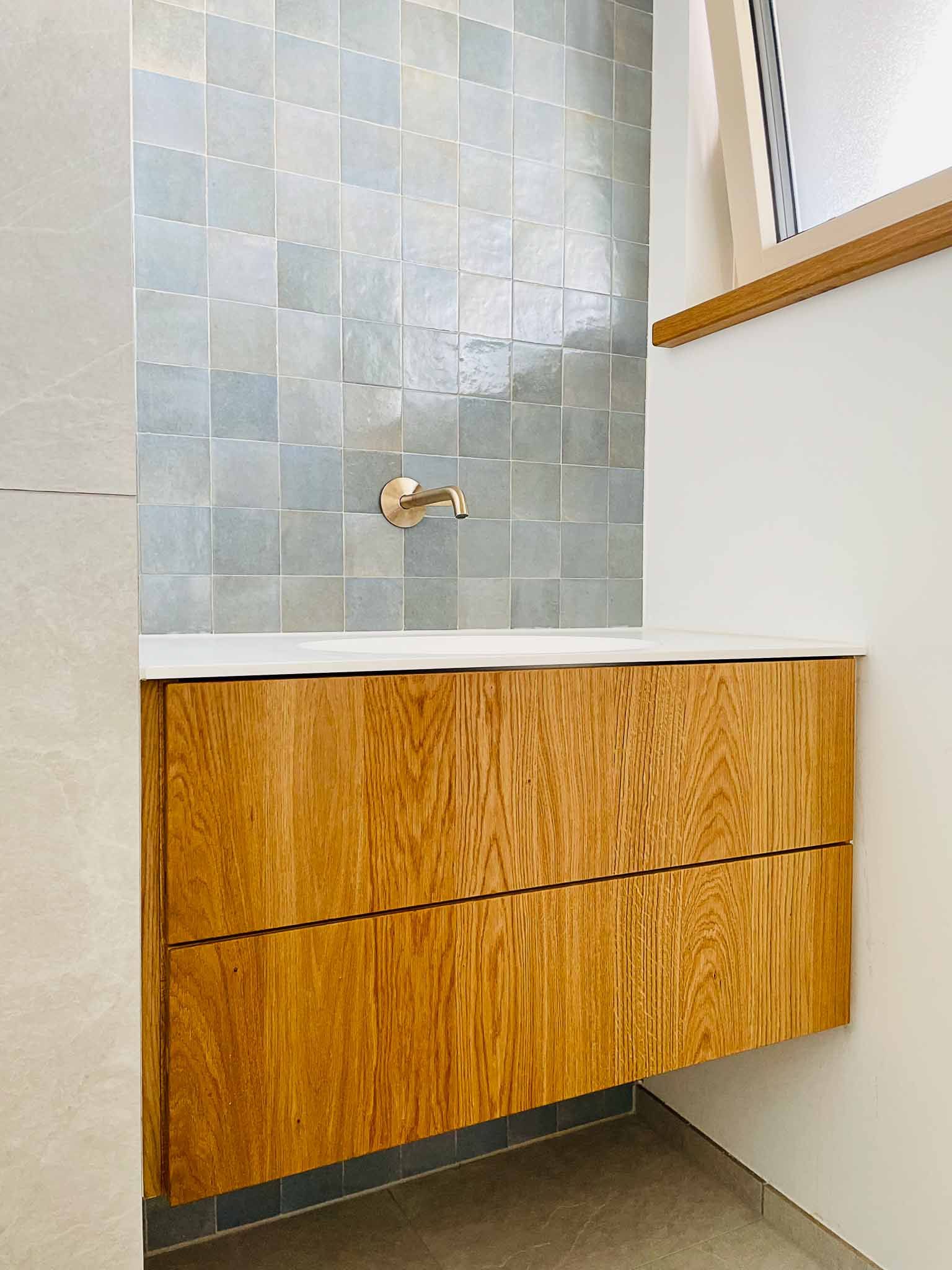 Schwebender Waschtisch aus Holz mit zwei Schubladen und einem goldenen Wasserhahn unter einer blauen quadratischen Fliesenrückwand.