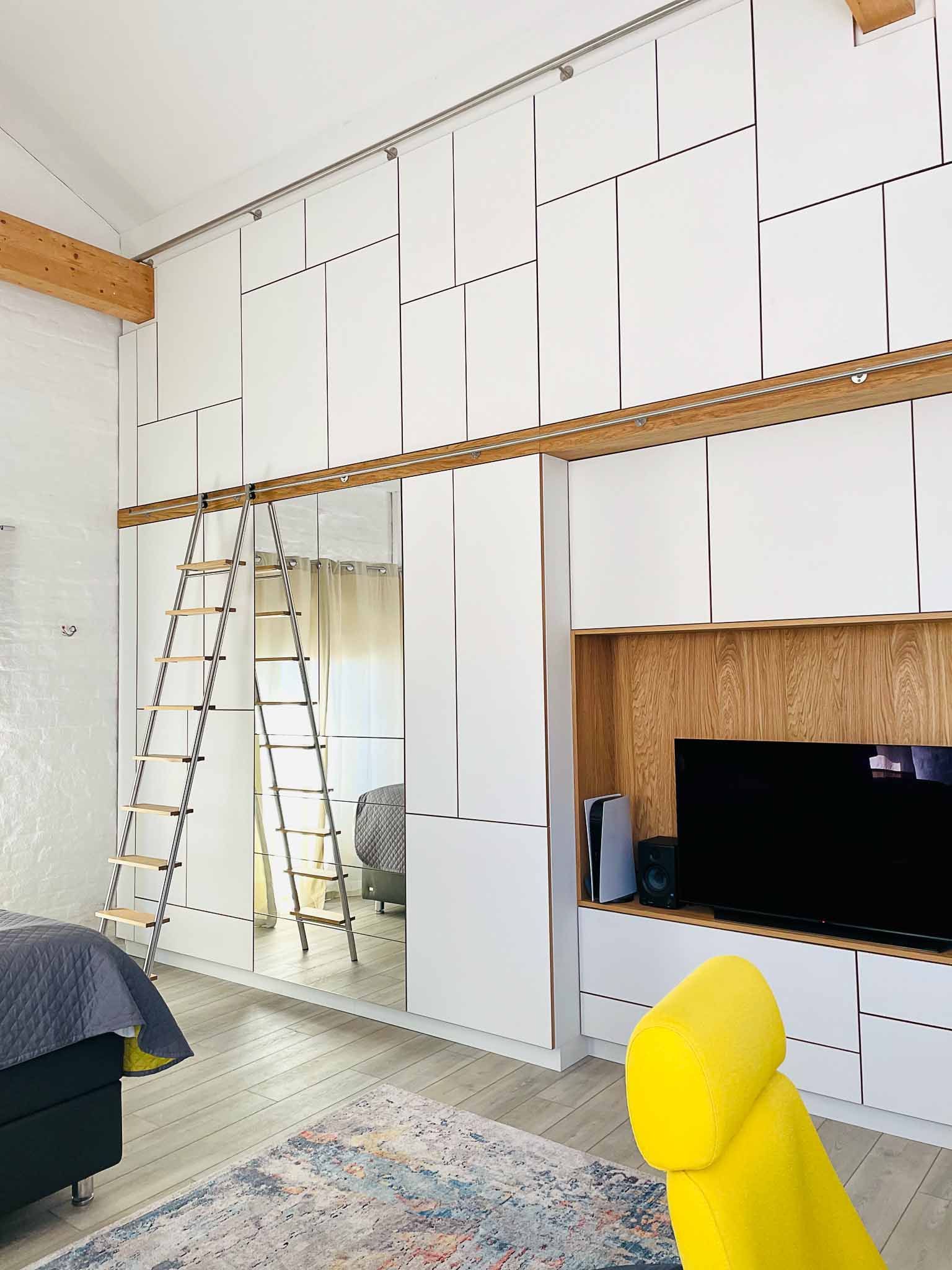 Loft-Schlafzimmer mit weißen Paneelwänden, Holzbalken und einer Leiter; im Vordergrund steht ein gelber Stuhl.