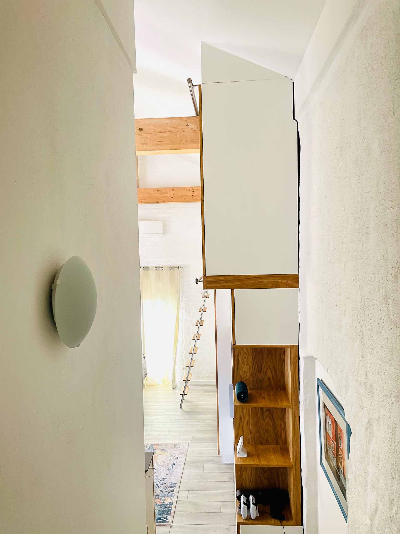 Schmaler Flur mit weißen Wänden, eingebauten Holzschränken und Blick in einen lichtdurchfluteten Raum mit einer Leiter.