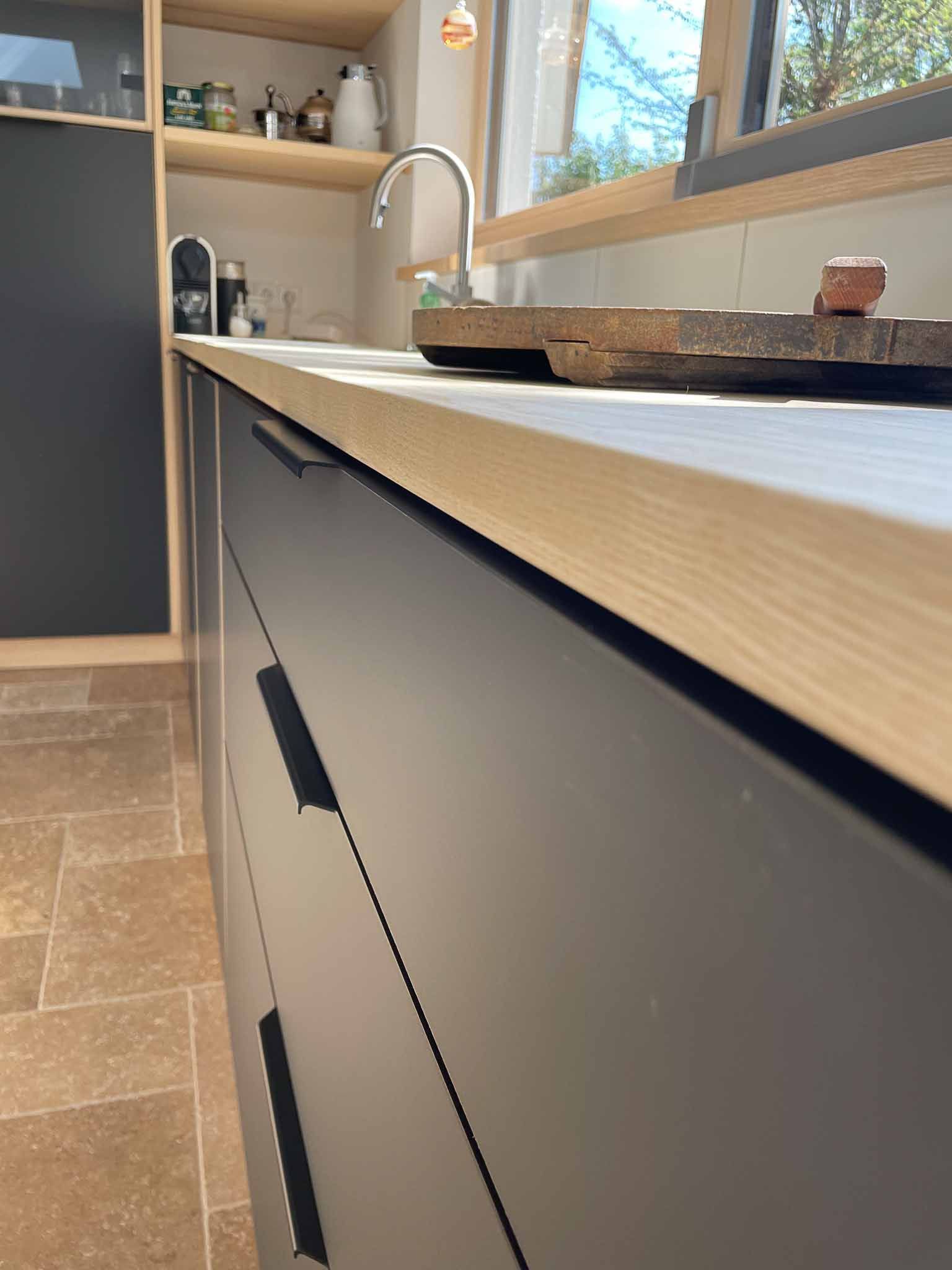 Graue Küchenschränke mit schwarzen Griffen, Arbeitsplatte, Spüle und offenen Regalen aus hellem Holz.