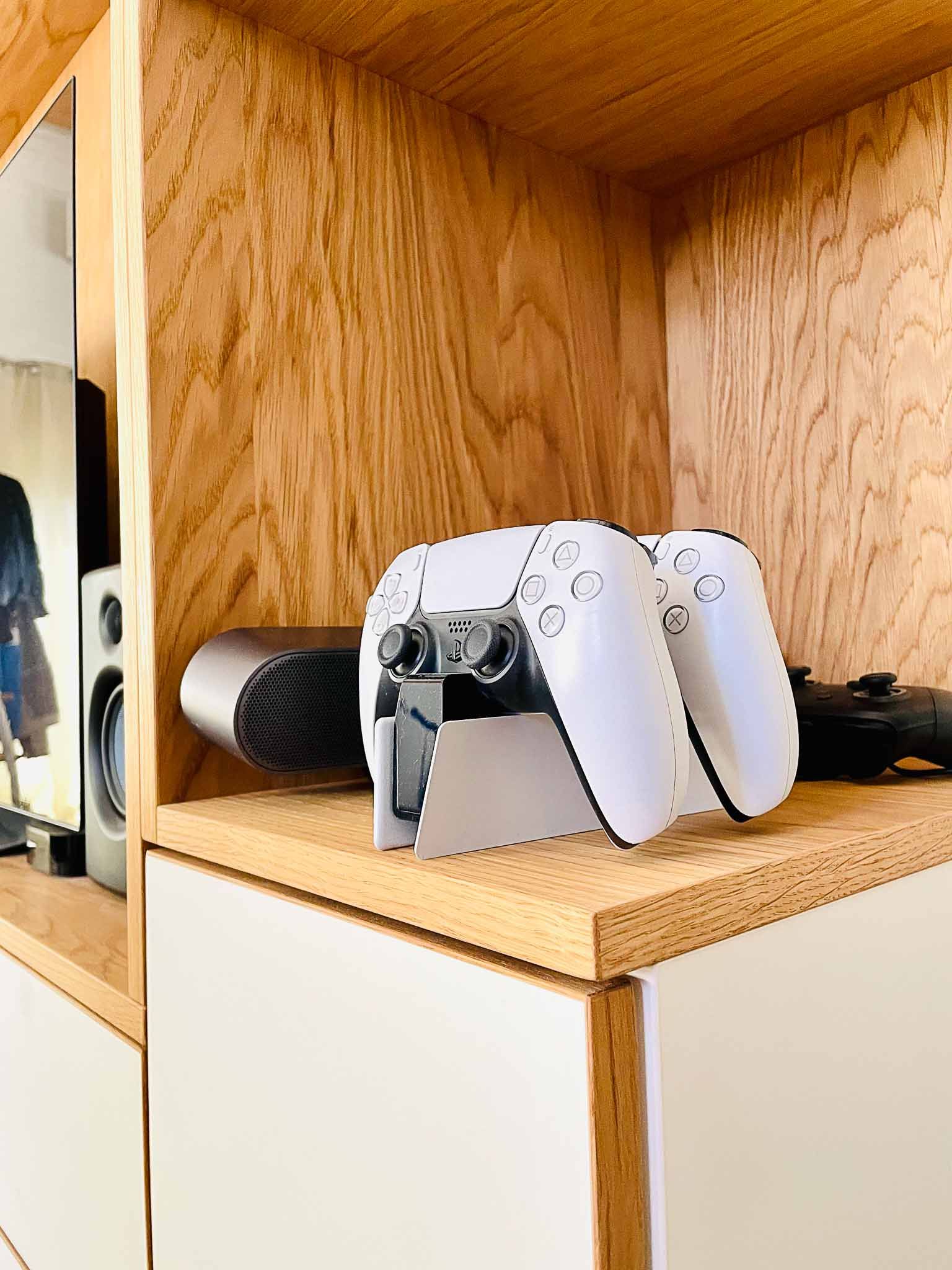 Zwei weiße PlayStation 5-Controller auf einem Regal in einem Holzschrank, mit einem Lautsprecher und anderen Gamecontrollern.