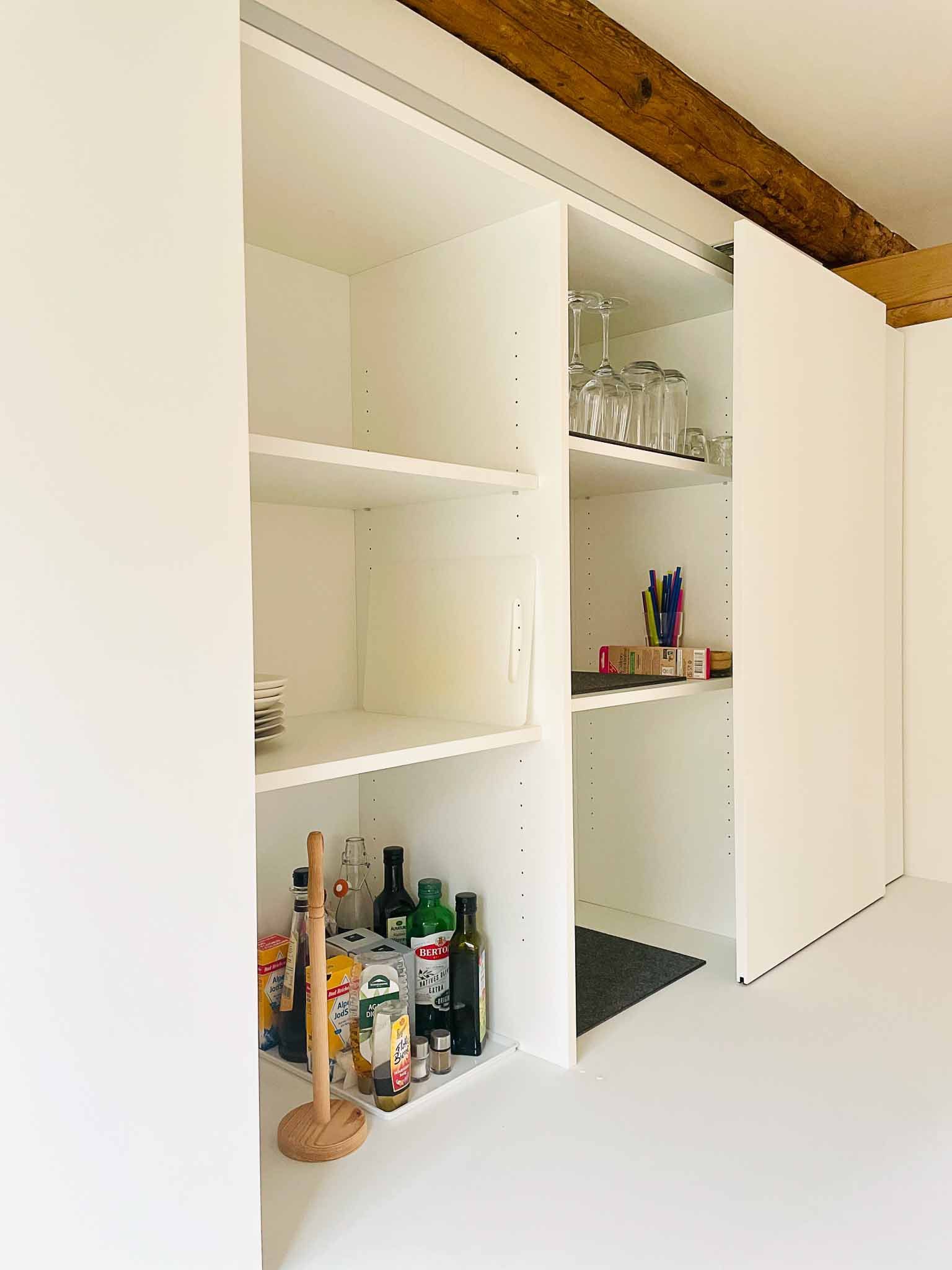 Weiße Speisekammer mit Schiebetüren, die sich öffnen lassen und Regale mit Glaswaren und Lebensmitteln freigeben. Rustikaler Holzbalken an der Decke.