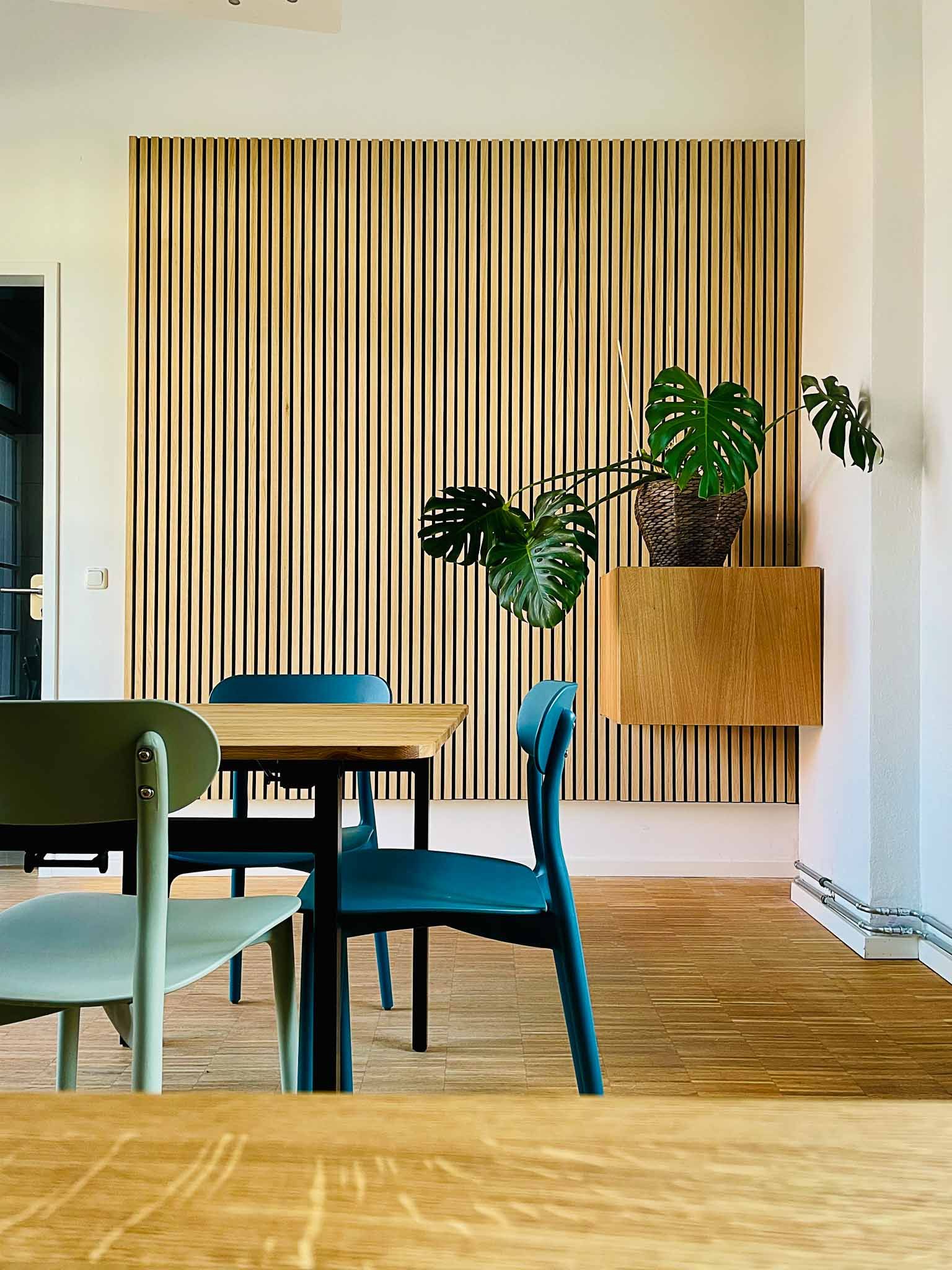 Essbereich mit Holzakzentwand, Tisch und bunten Stühlen; Pflanze auf schwebendem Regal.