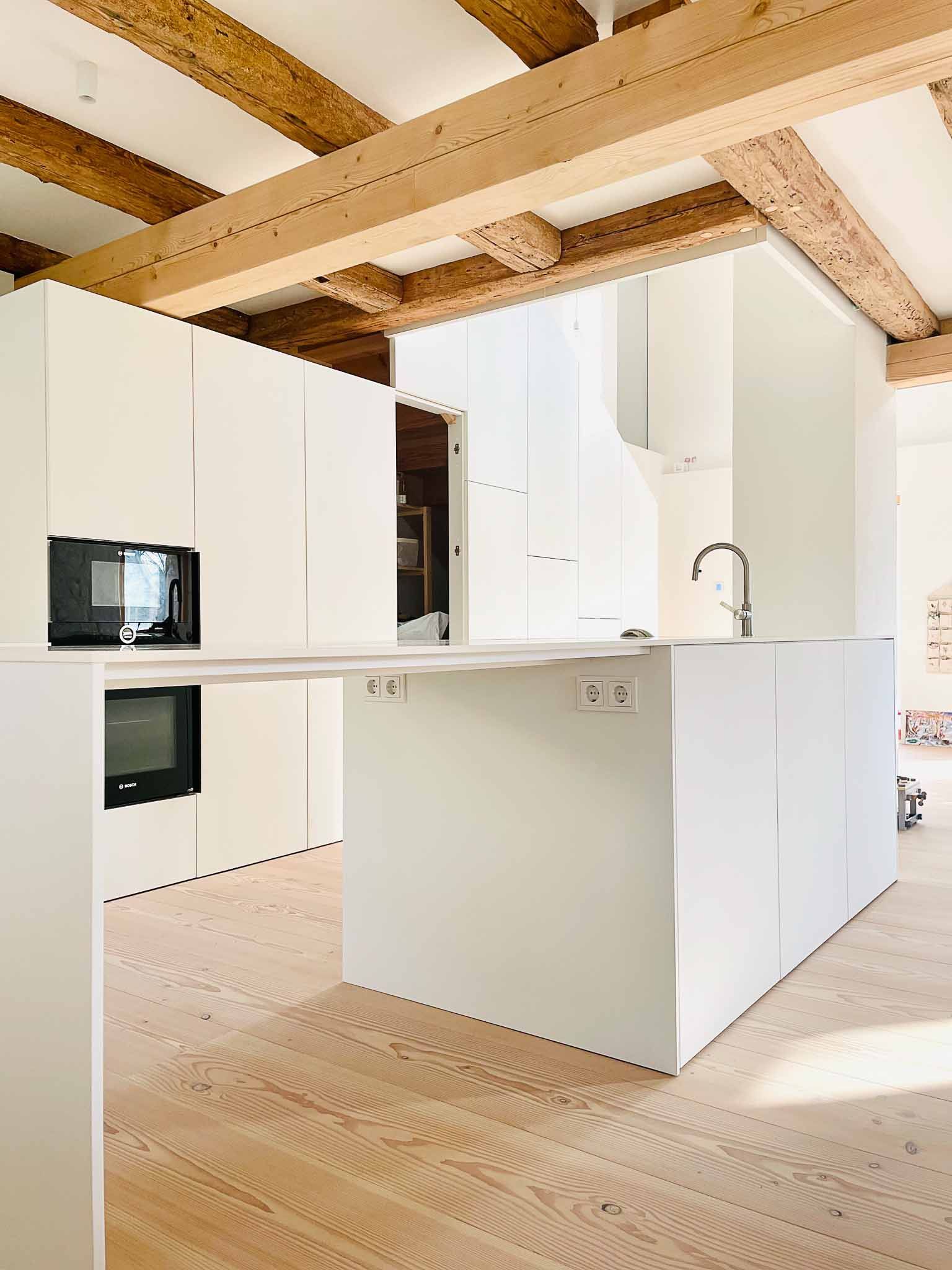 Weiße, moderne Küche mit Holzbalken, Kochinsel, Schränken und Geräten.