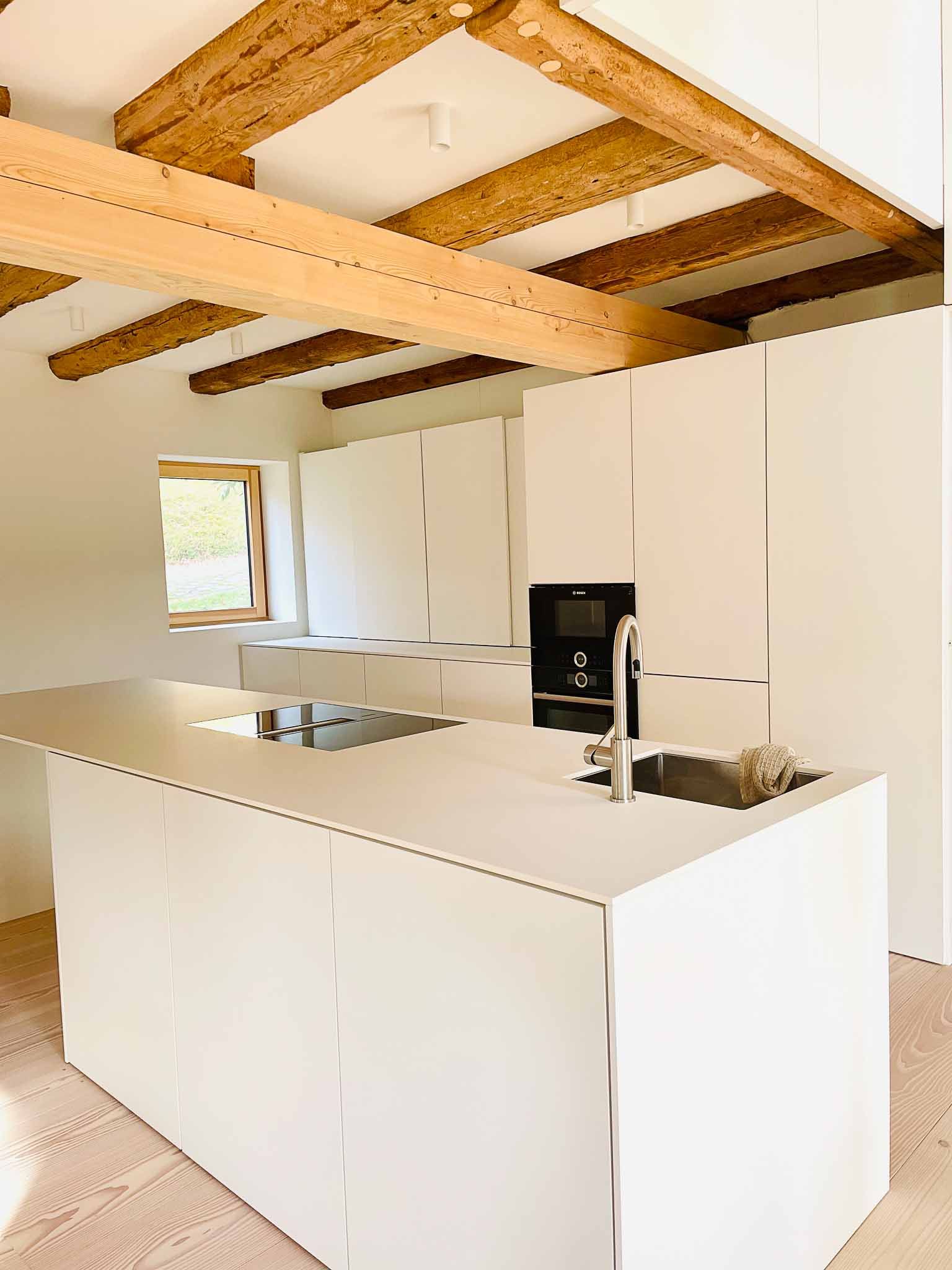 Moderne weiße Küche mit Kochinsel, Holzbalken und eingebauter Kaffeemaschine.