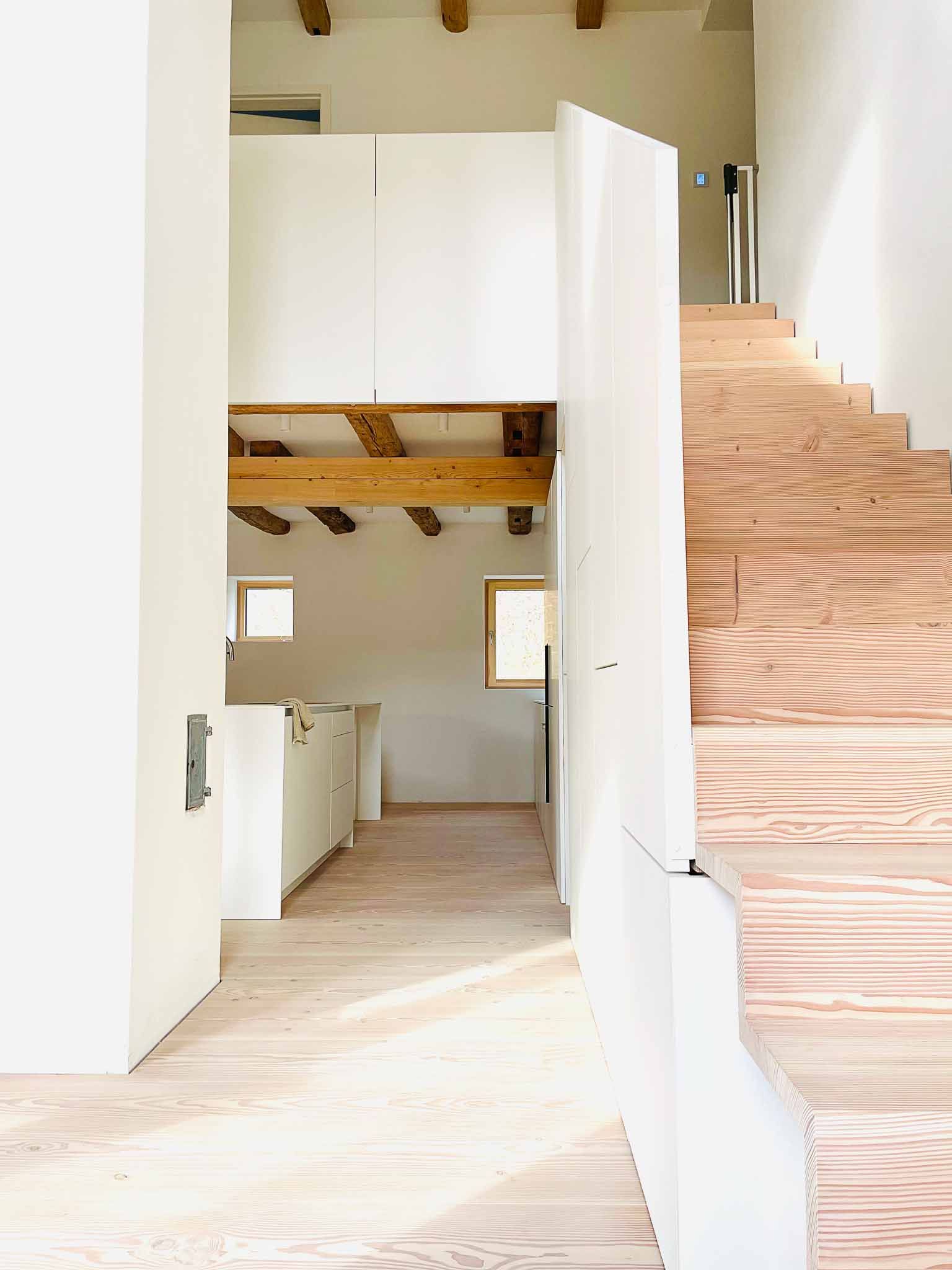 Innenansicht mit Holztreppe und Blick auf eine Küche mit weißen Schränken.