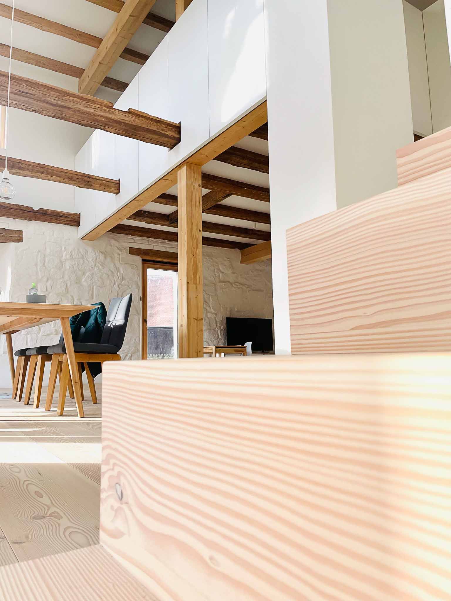 Eine Holztreppe führt hinauf in einen lichtdurchfluteten Innenraum mit freiliegenden Balken, einem Esstisch und einem Fenster.