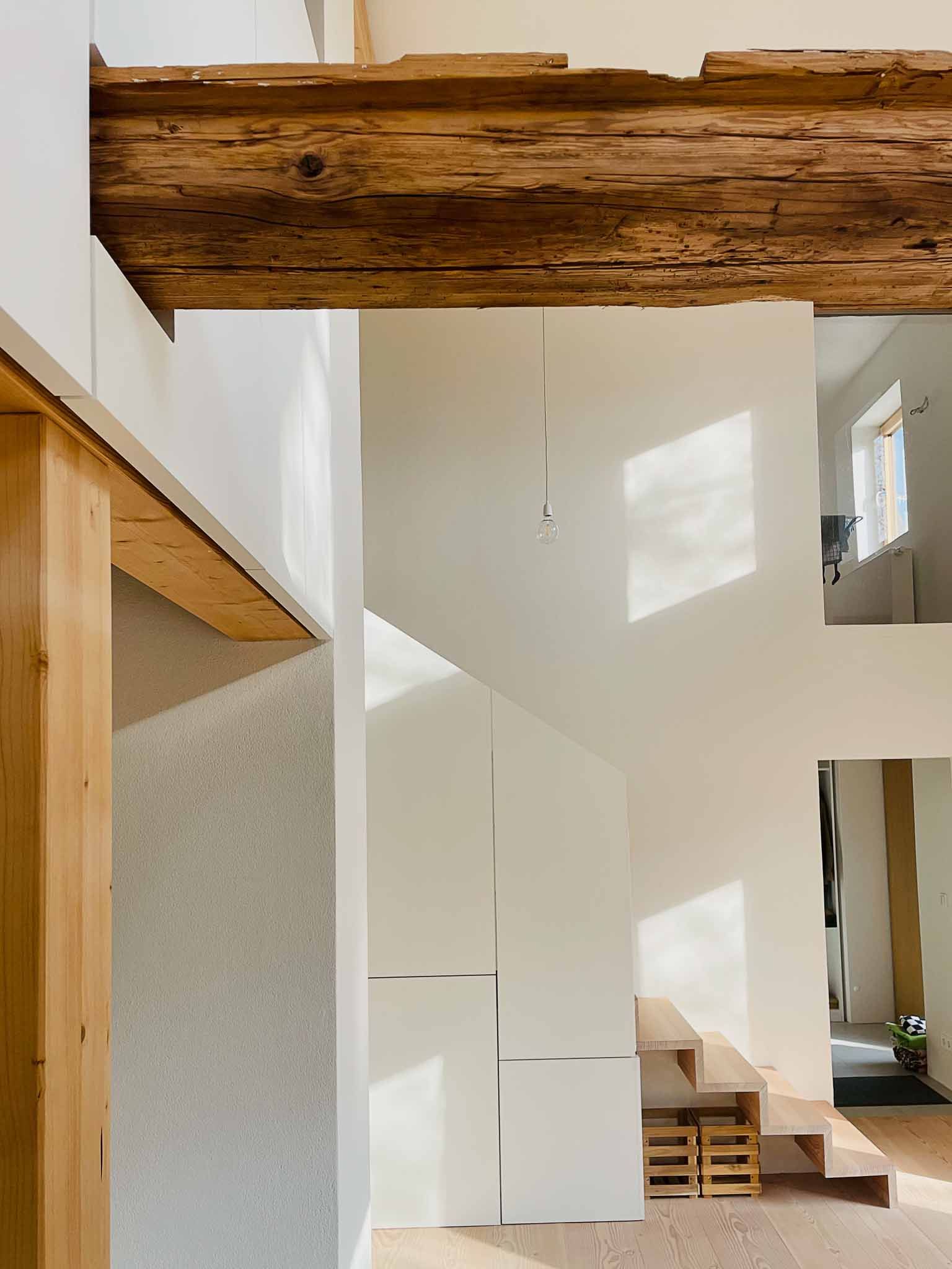 Helles Interieur mit Holzbalken, weißen Wänden und einer vom Sonnenlicht beleuchteten Treppe.