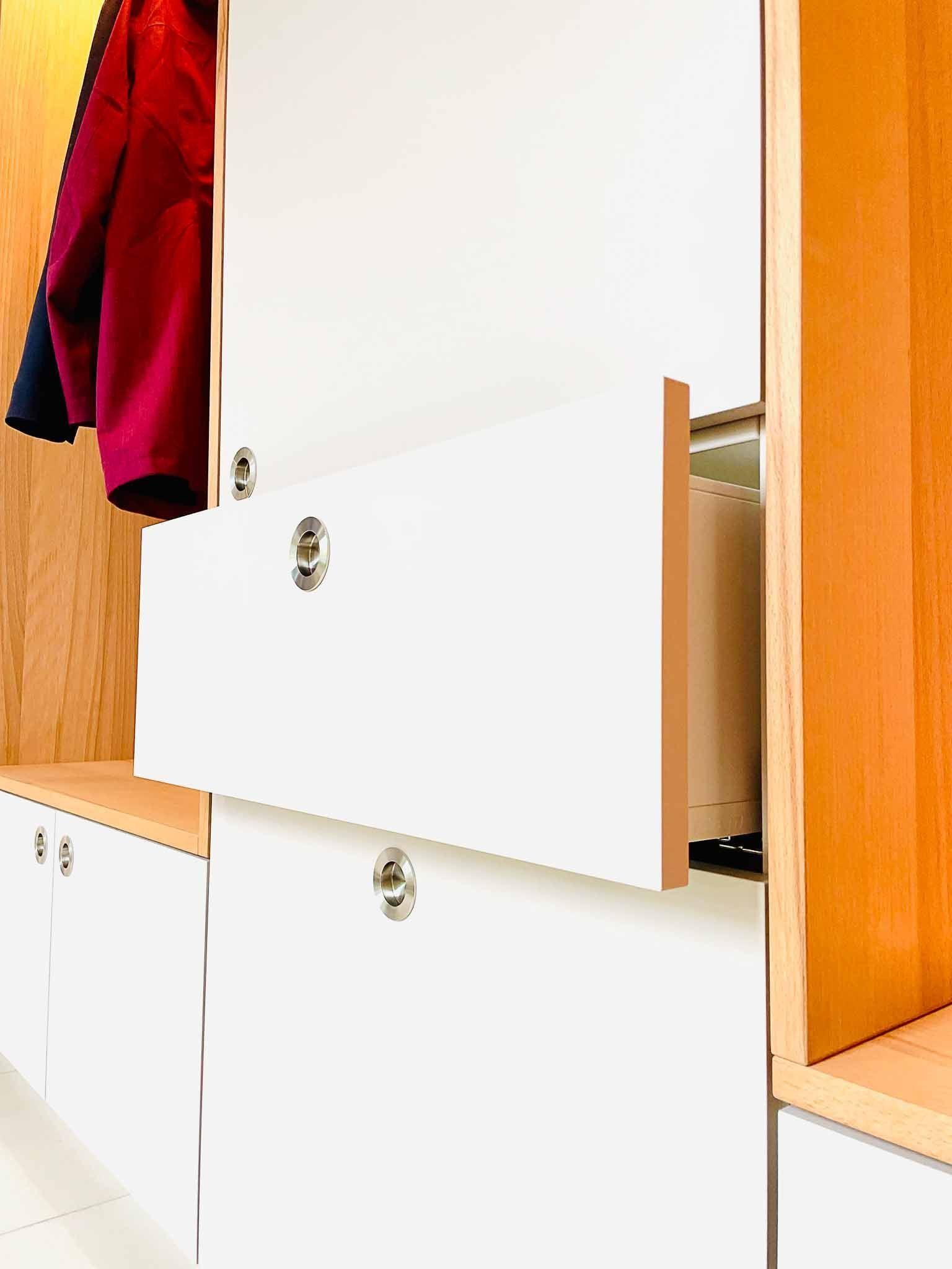 Weißer Schrank mit einer offenen Schublade, die den Blick auf das Innere freigibt und an der ein roter Mantel hängt.