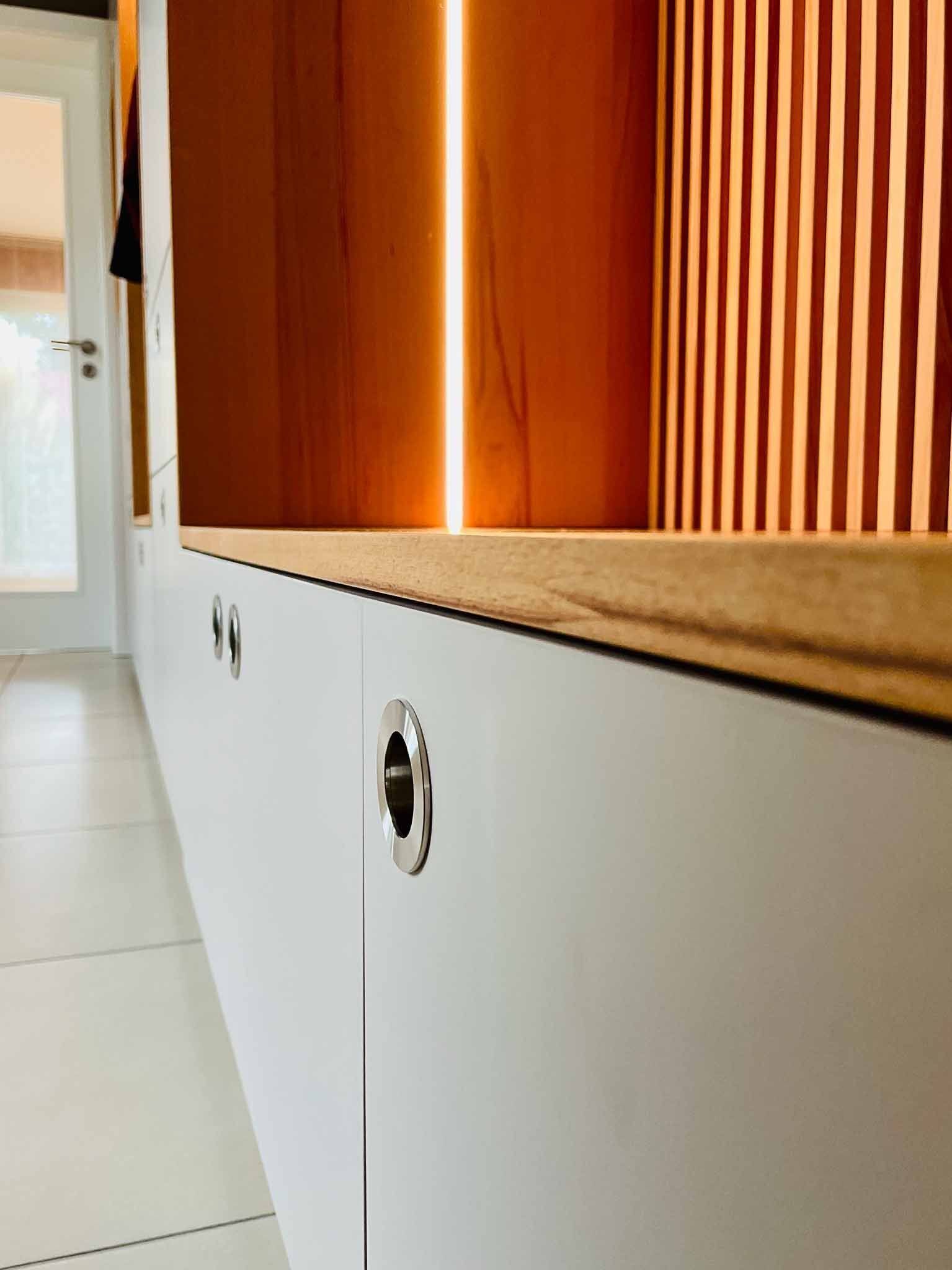 Weißer Schrank mit Metallgriffen, Holzregal, orange hinterleuchteter Wand und vertikalen Holzpaneelen.