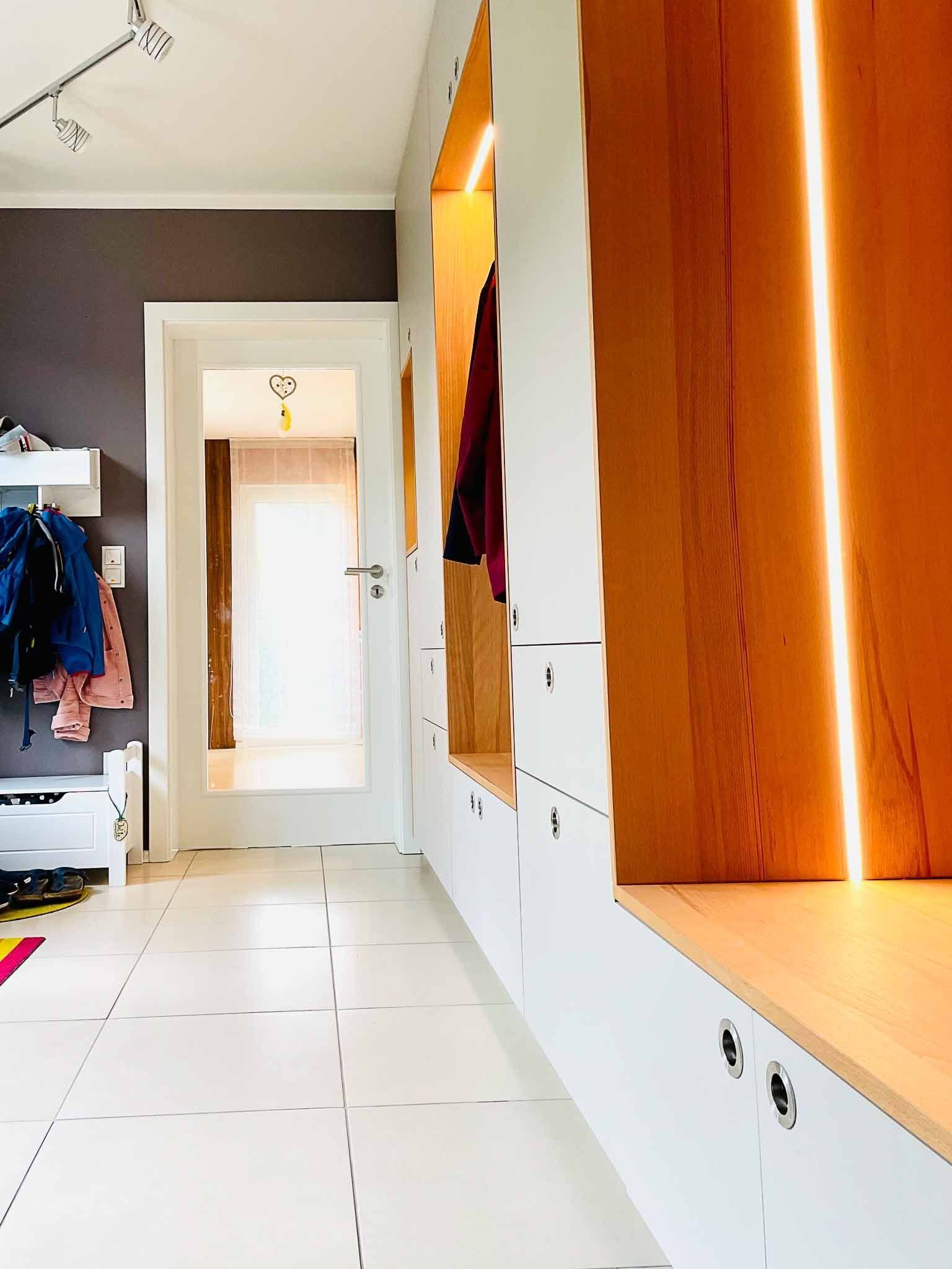 Flur mit weißem Boden und eingebautem Stauraum. Holzverkleidung mit warmer Beleuchtung, eine Tür am Ende.