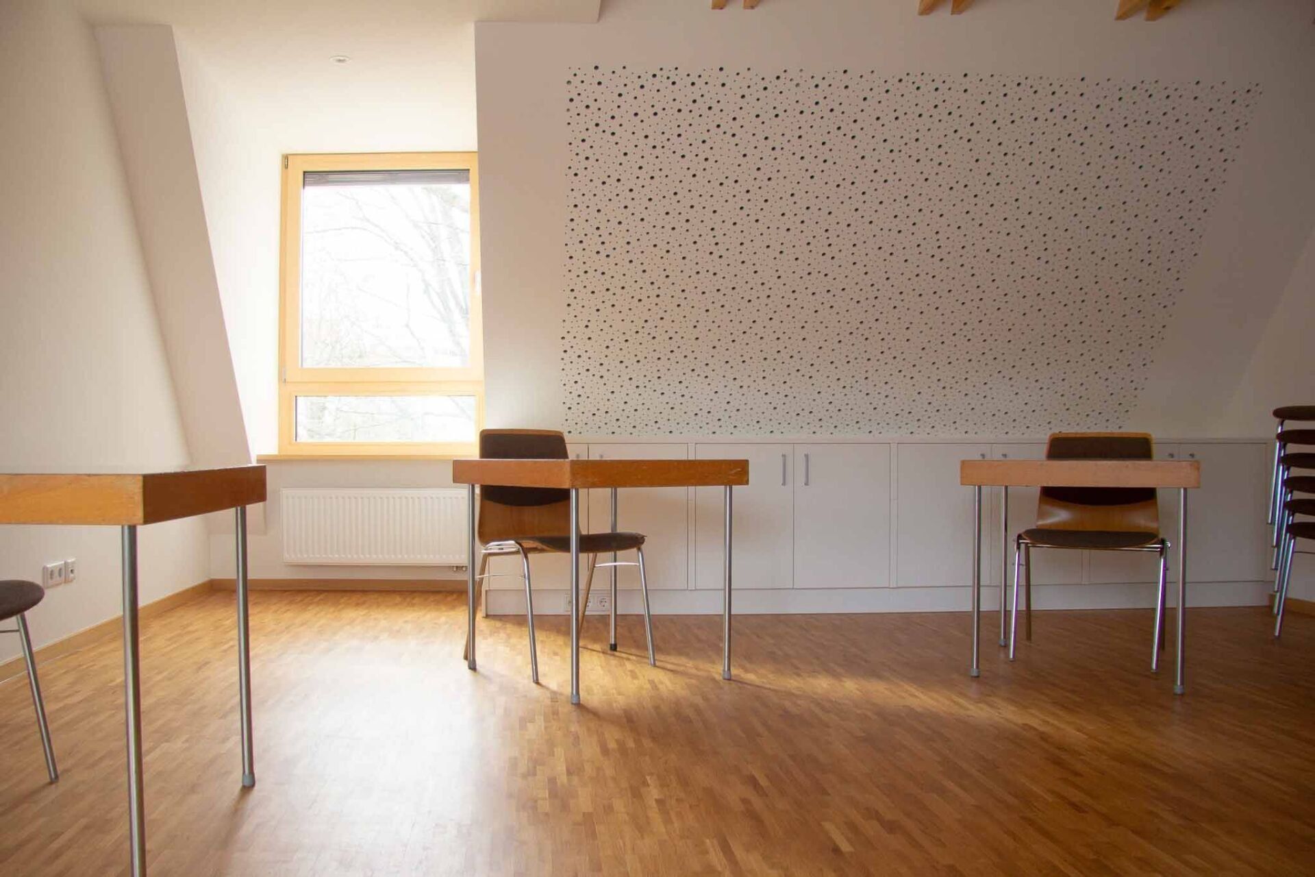 Leeres Klassenzimmer mit Holztischen und -stühlen, einem Fenster und einer Wand mit gepunktetem Muster.