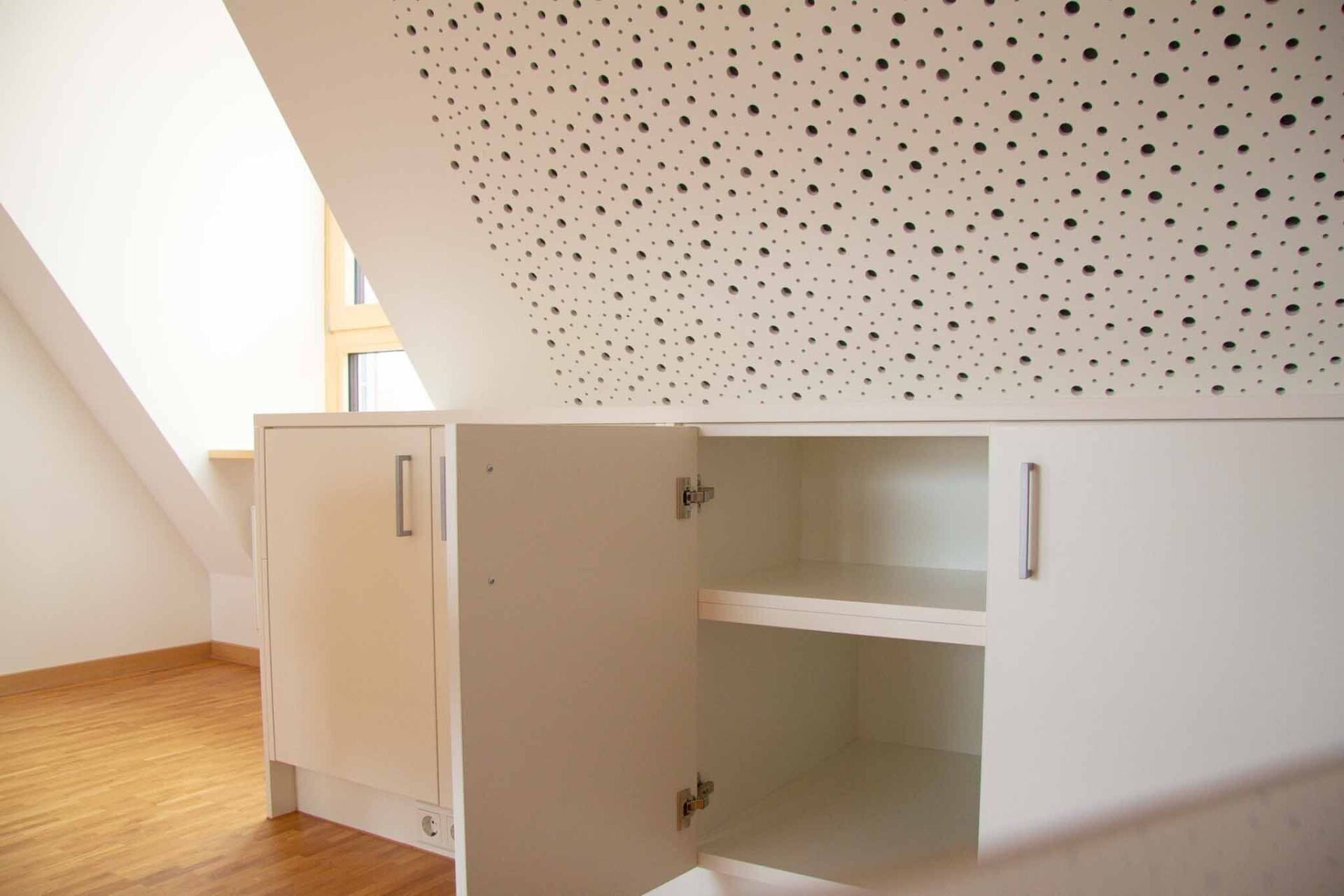 Weiße Schränke mit einer offenen Tür in einem Raum mit Dachschräge und Holzboden.