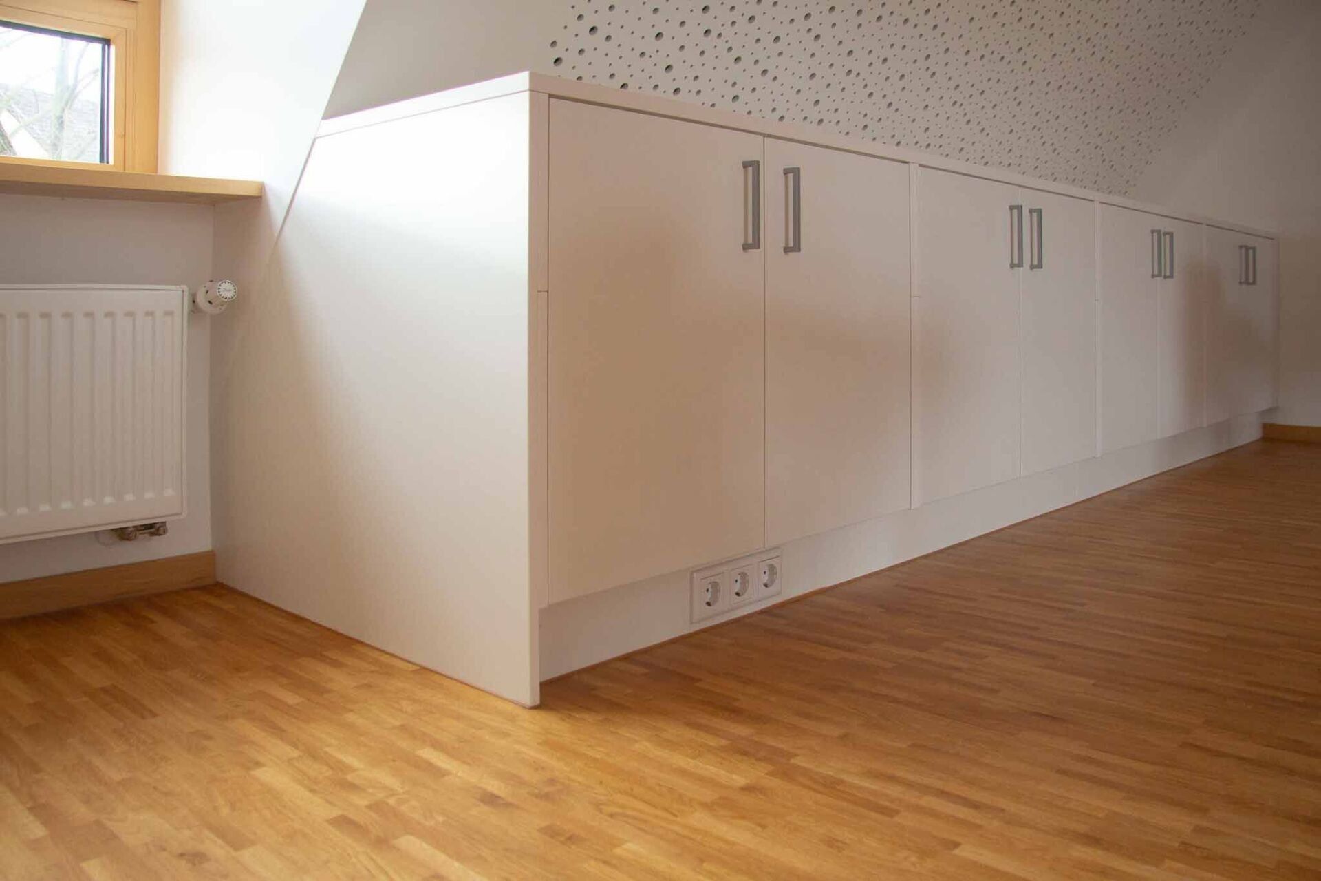 Eingebaute weiße Lagerschränke entlang einer Wand mit Hartholzboden, in der Nähe eines Heizkörpers und eines Fensters.