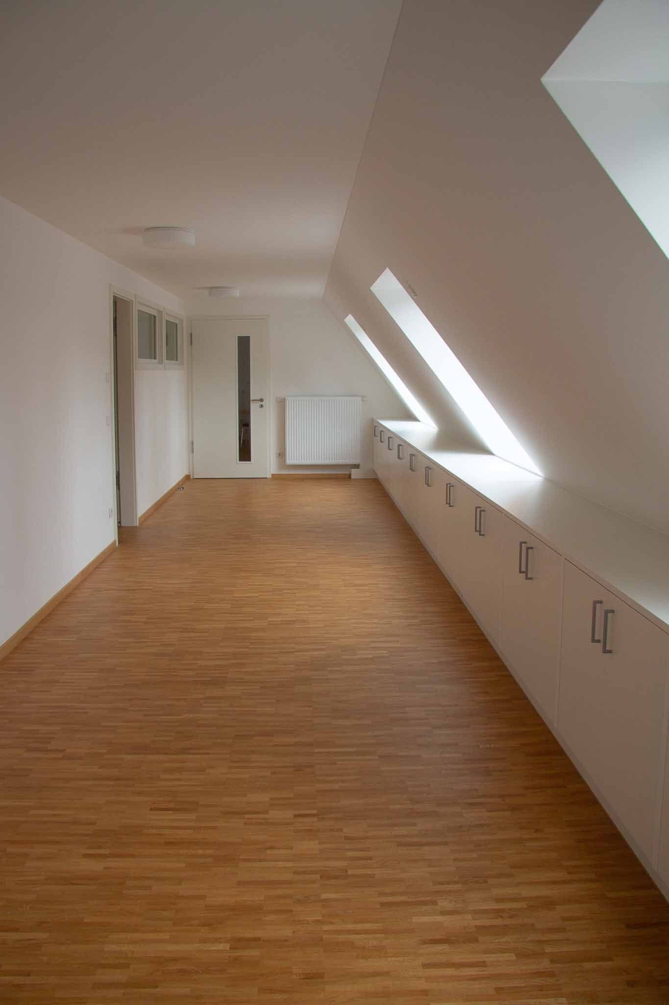 Leerer Dachbodenraum mit Holzboden, Dachschräge, weißen Wänden, Einbauschränken und kleinen Fenstern.
