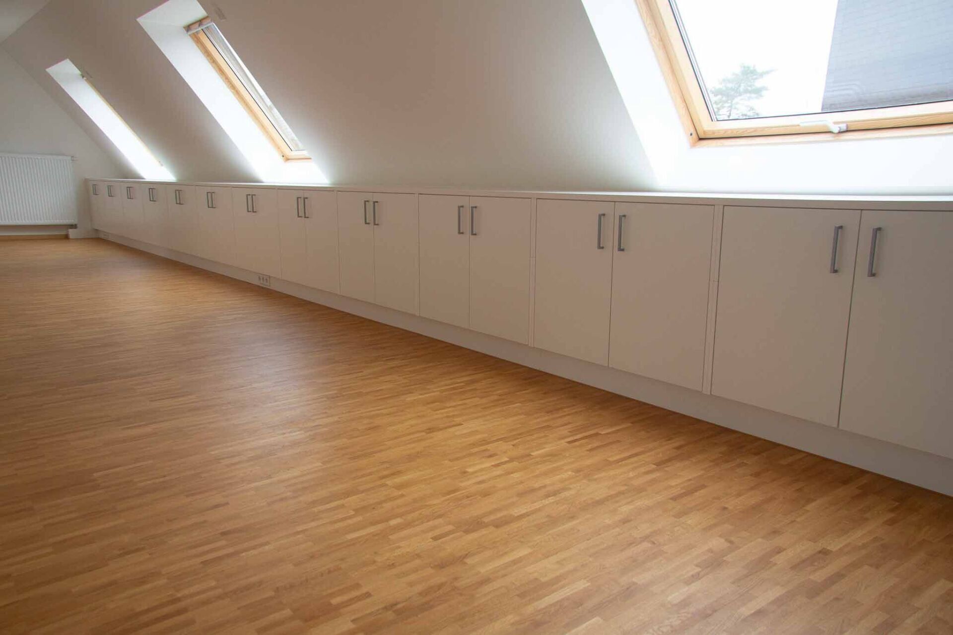 Dachzimmer mit Holzboden und einer langen Reihe weißer Lagerschränke unter schrägen Oberlichtern.