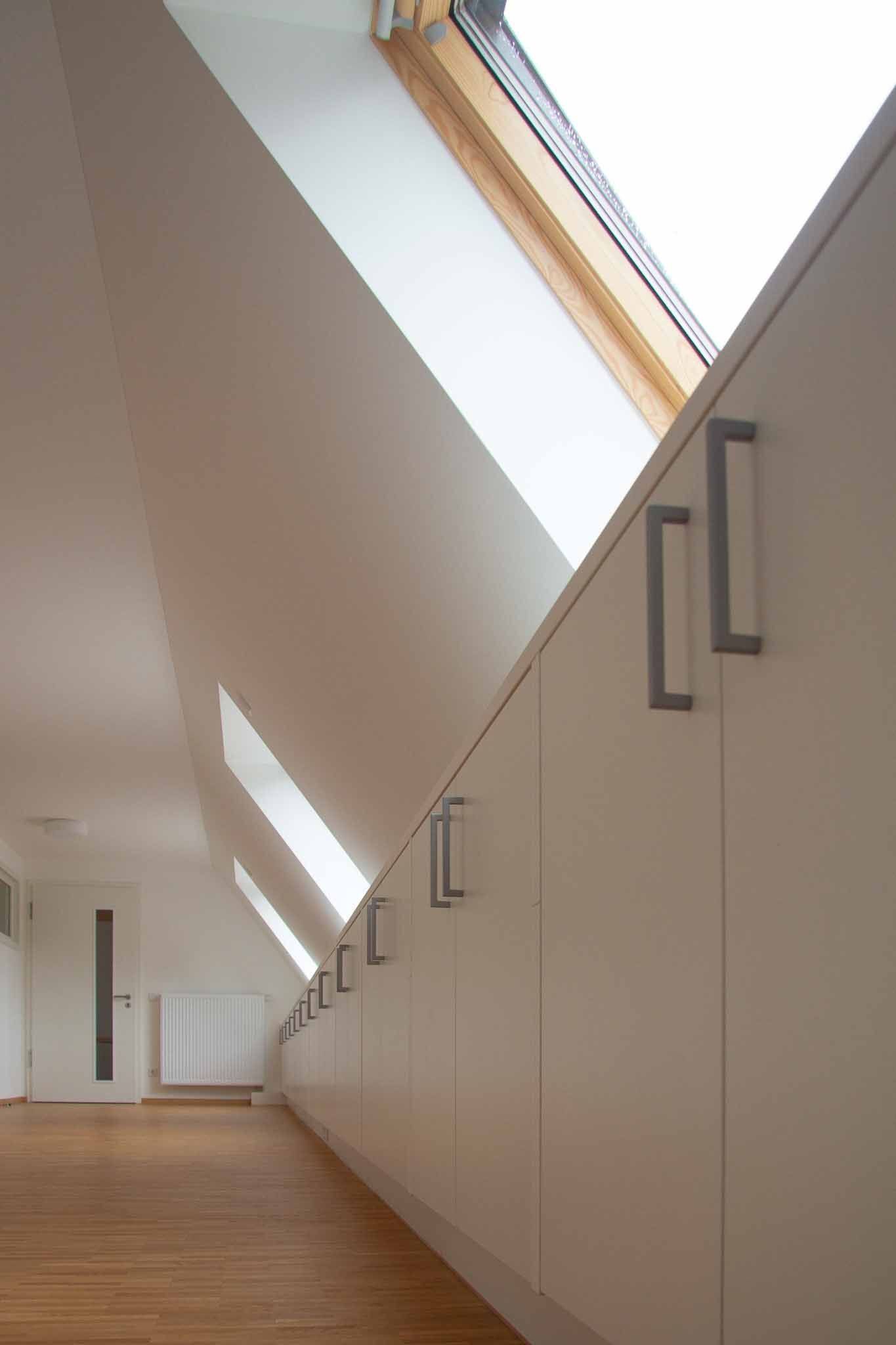 Weiße Einbauschränke entlang einer Dachschräge mit Oberlicht. Holzboden, Heizkörper und Tür sichtbar.