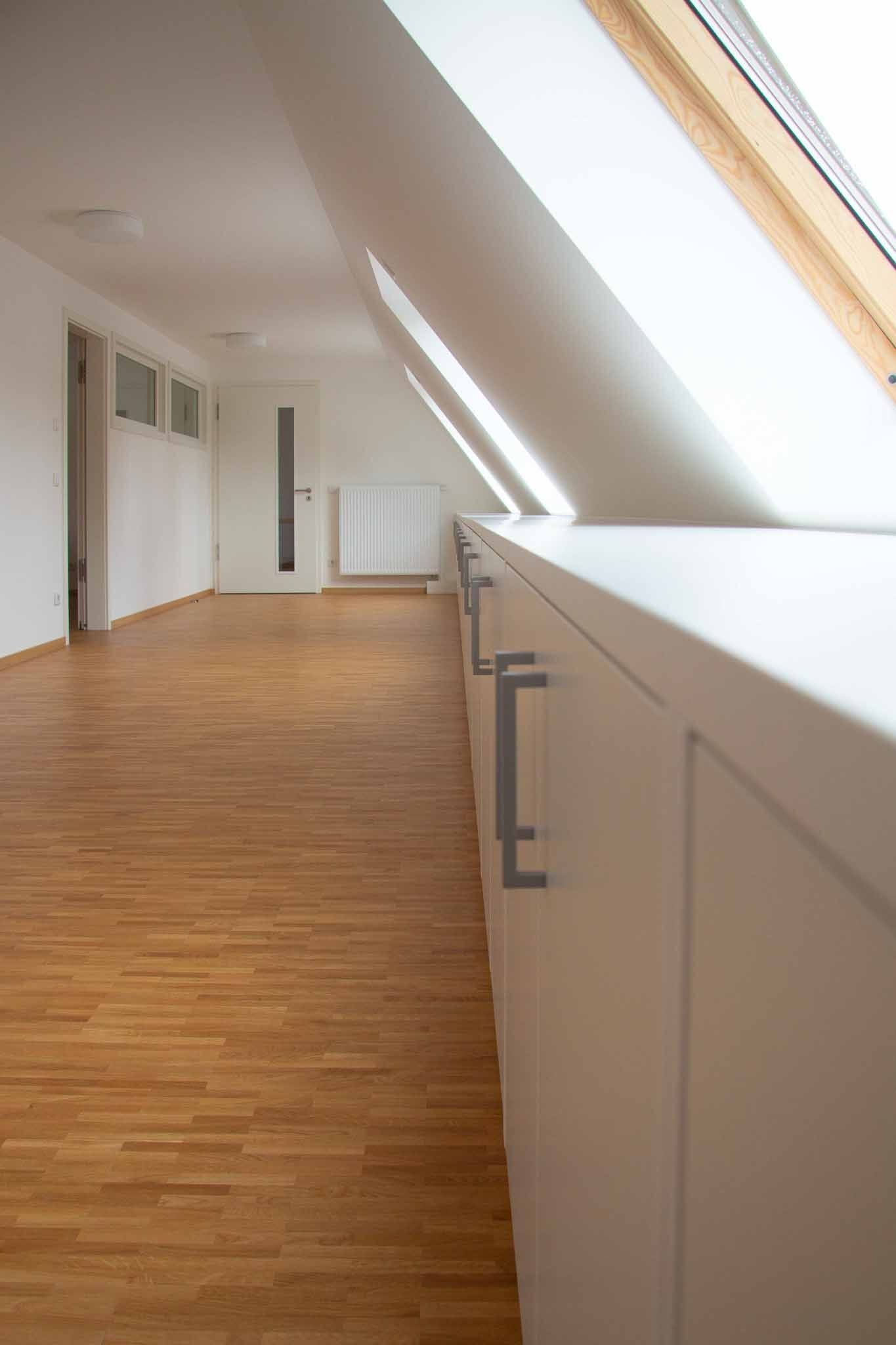 Langer, heller Raum mit Holzboden, eingebauten weißen Schränken und schrägen Oberlichtern.