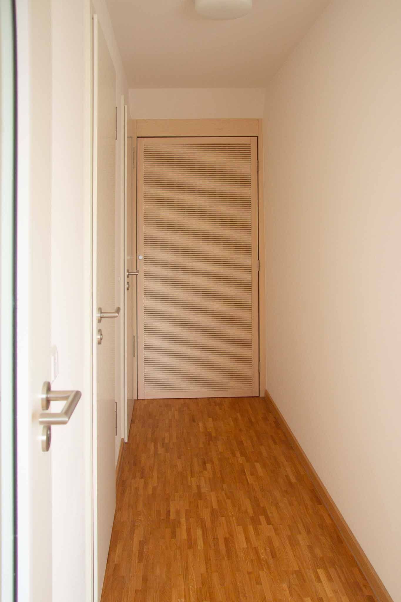 Flur mit Holzboden, hellen Wänden und einer Tür mit diagonalen Akzenten aus Milchglas.