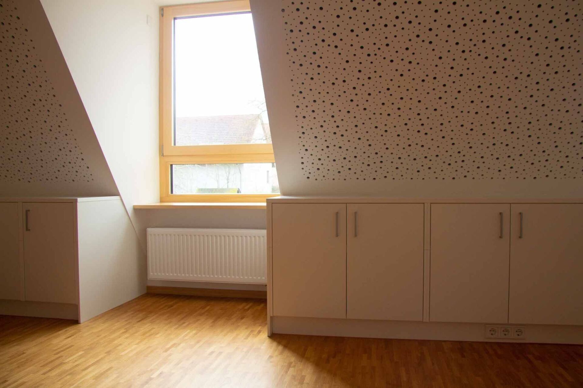 Ein Dachzimmer mit Dachschrägen, Fenster, Schränken und Holzboden. Die Decke weist eine perforierte Struktur auf.