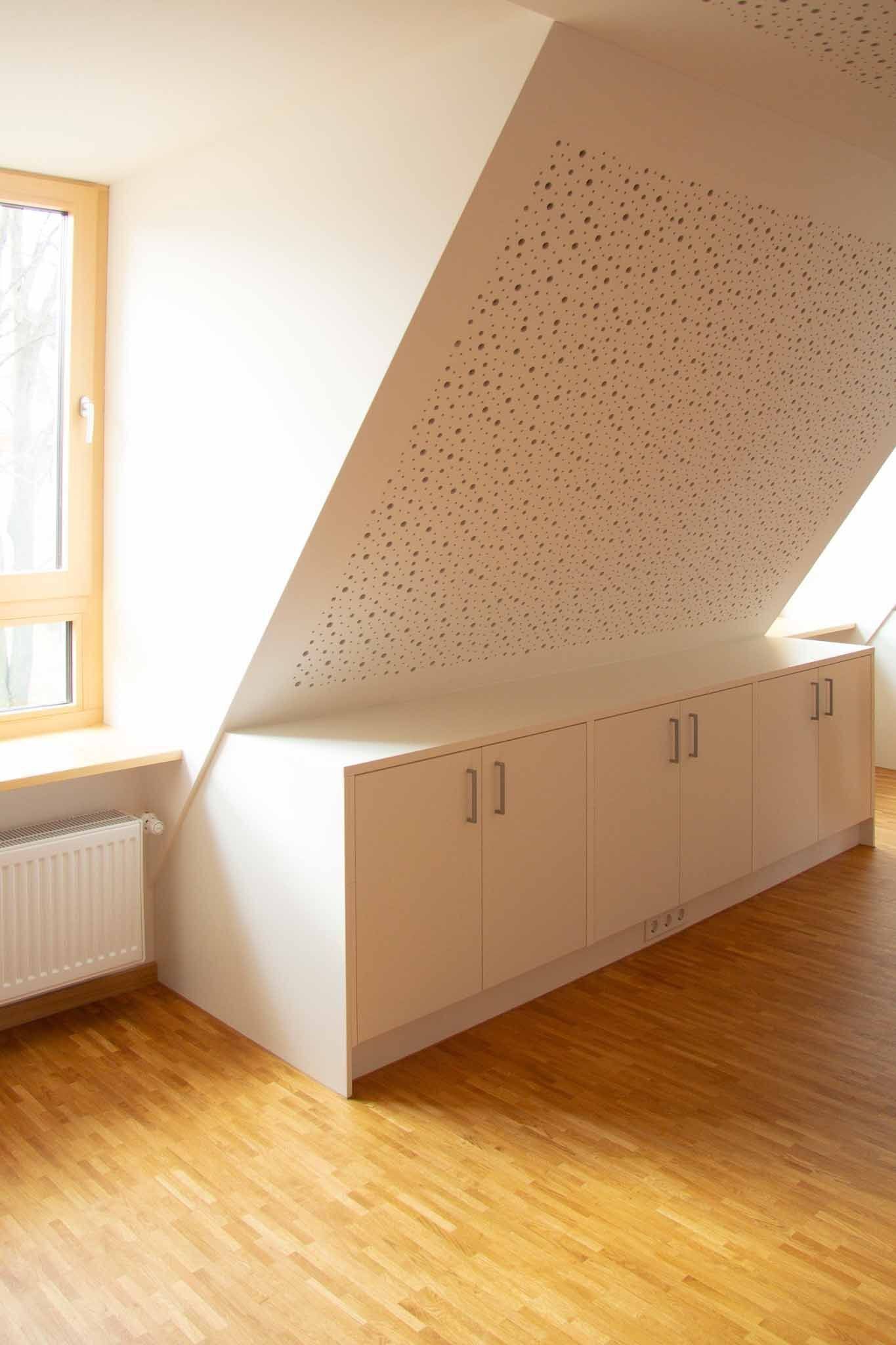 Weiße Einbauschränke unter einer schrägen, perforierten Decke. Fenster und Heizkörper links, Holzboden.