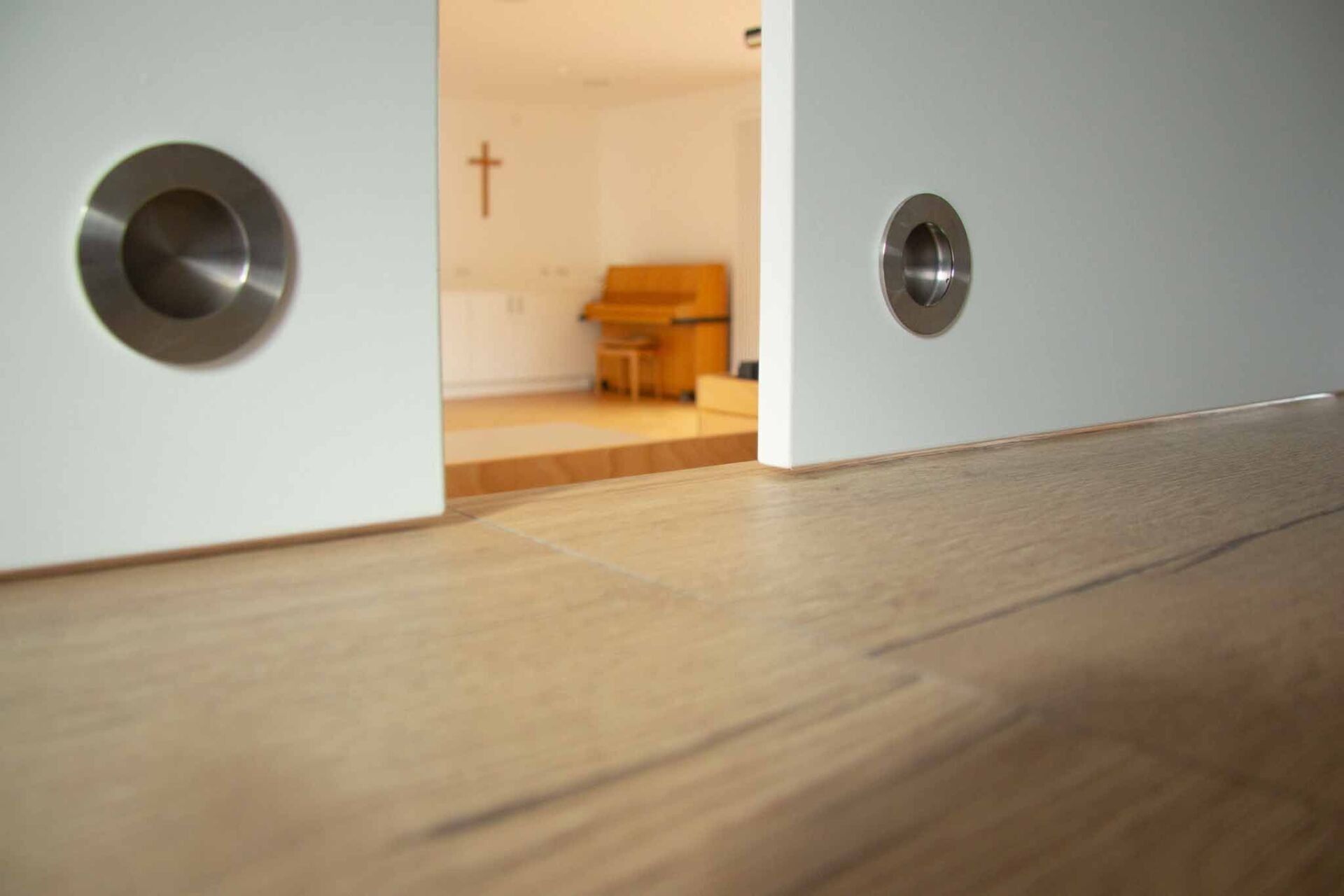 Weiße Schiebetüren mit runden Griffen öffnen sich und geben den Blick auf einen Raum frei, an dessen Wand ein Klavier und ein Kreuz hängen.