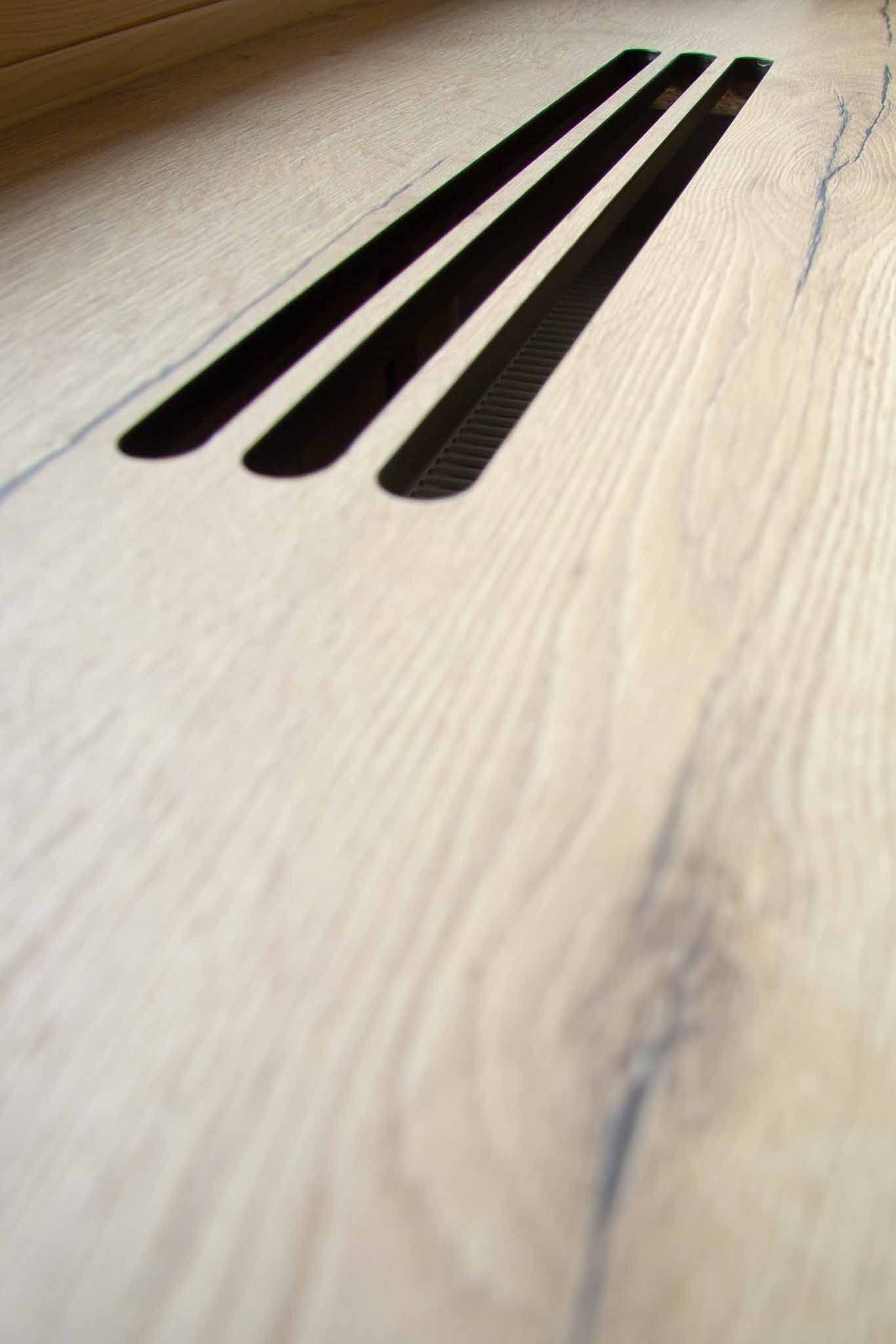 Drei dunkle, rechteckige Lüftungsschlitze in einem hellen Holzboden.