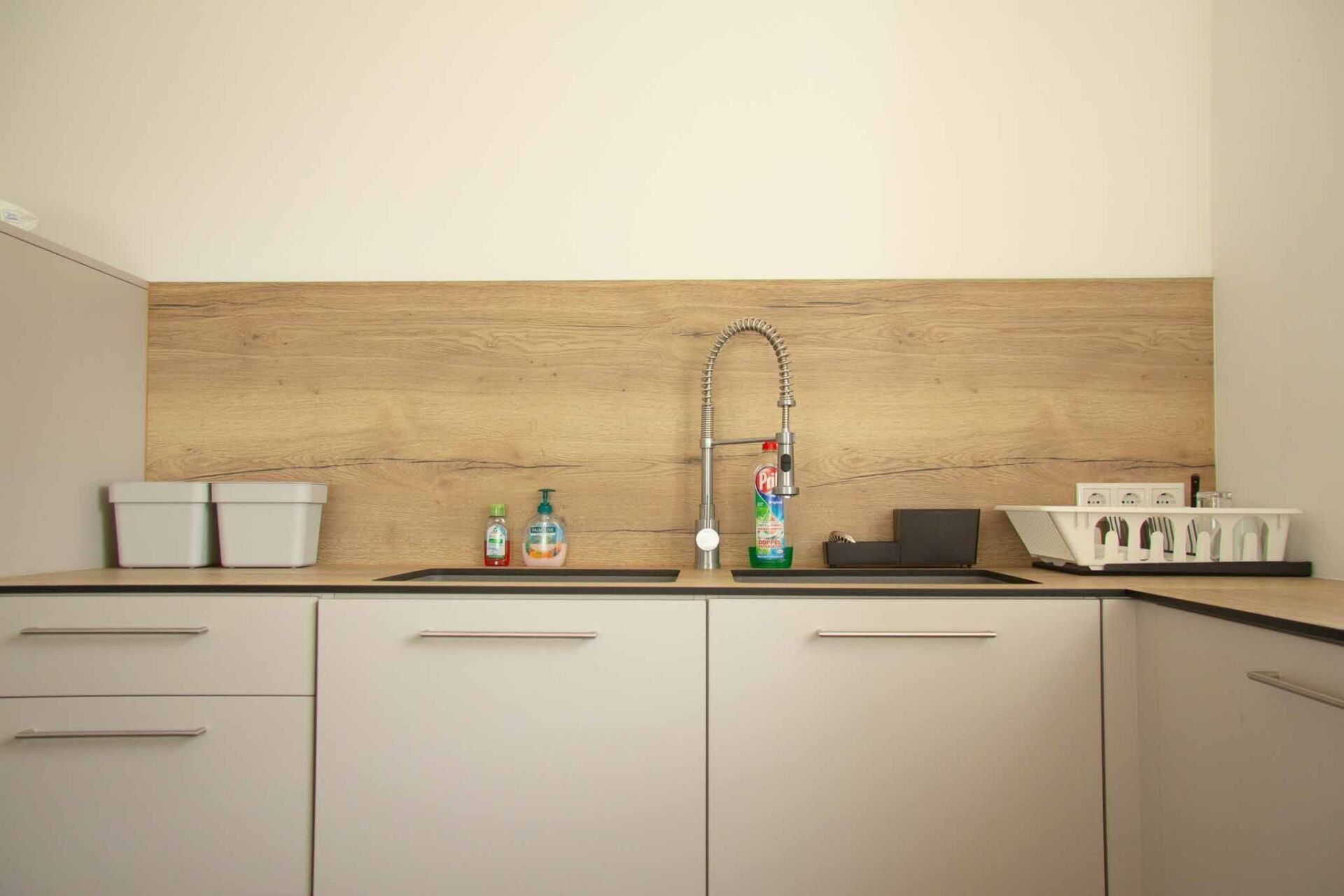 Küche mit hellen Schränken, Holzrückwand und einer Edelstahlspüle mit Reinigungsmitteln.