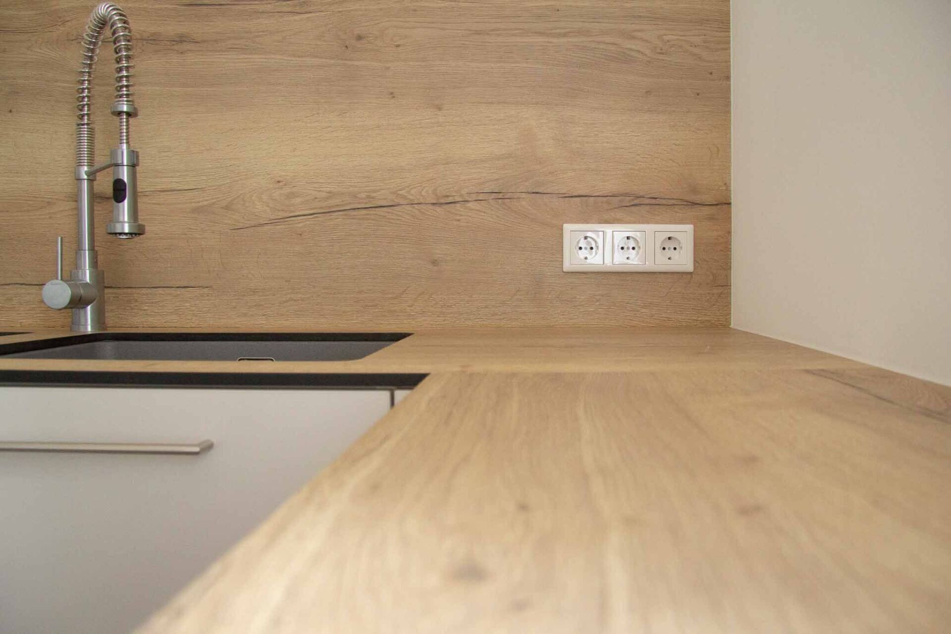 Küchentheke mit Holzoberfläche, Spüle, Wasserhahn und Steckdosen auf einer hellen Holzrückwand.