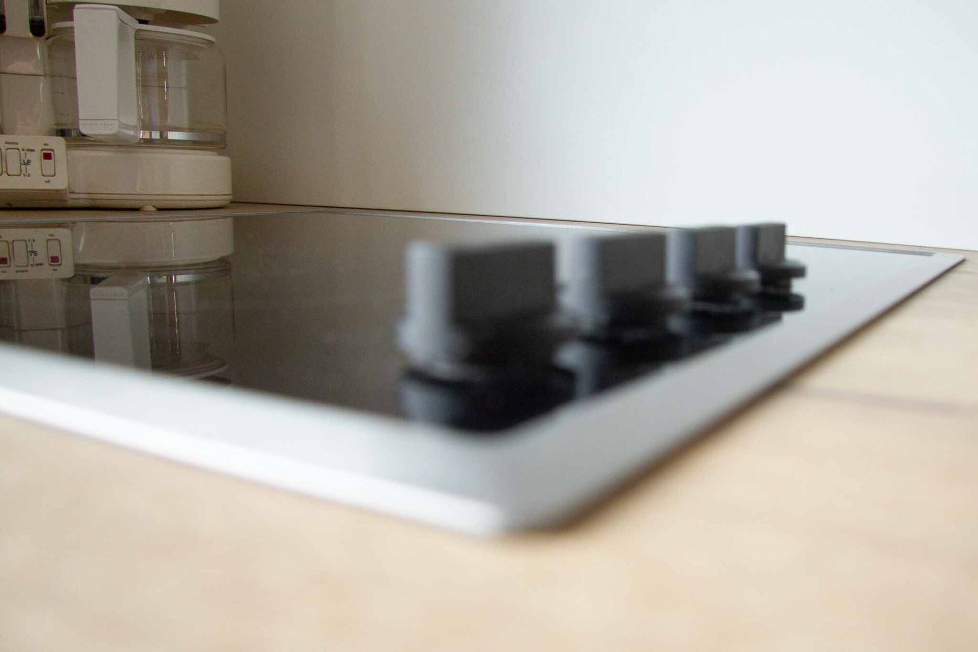 Eine schwarze Herdplatte mit vier Knöpfen. Im Hintergrund steht eine weiße Kaffeemaschine.