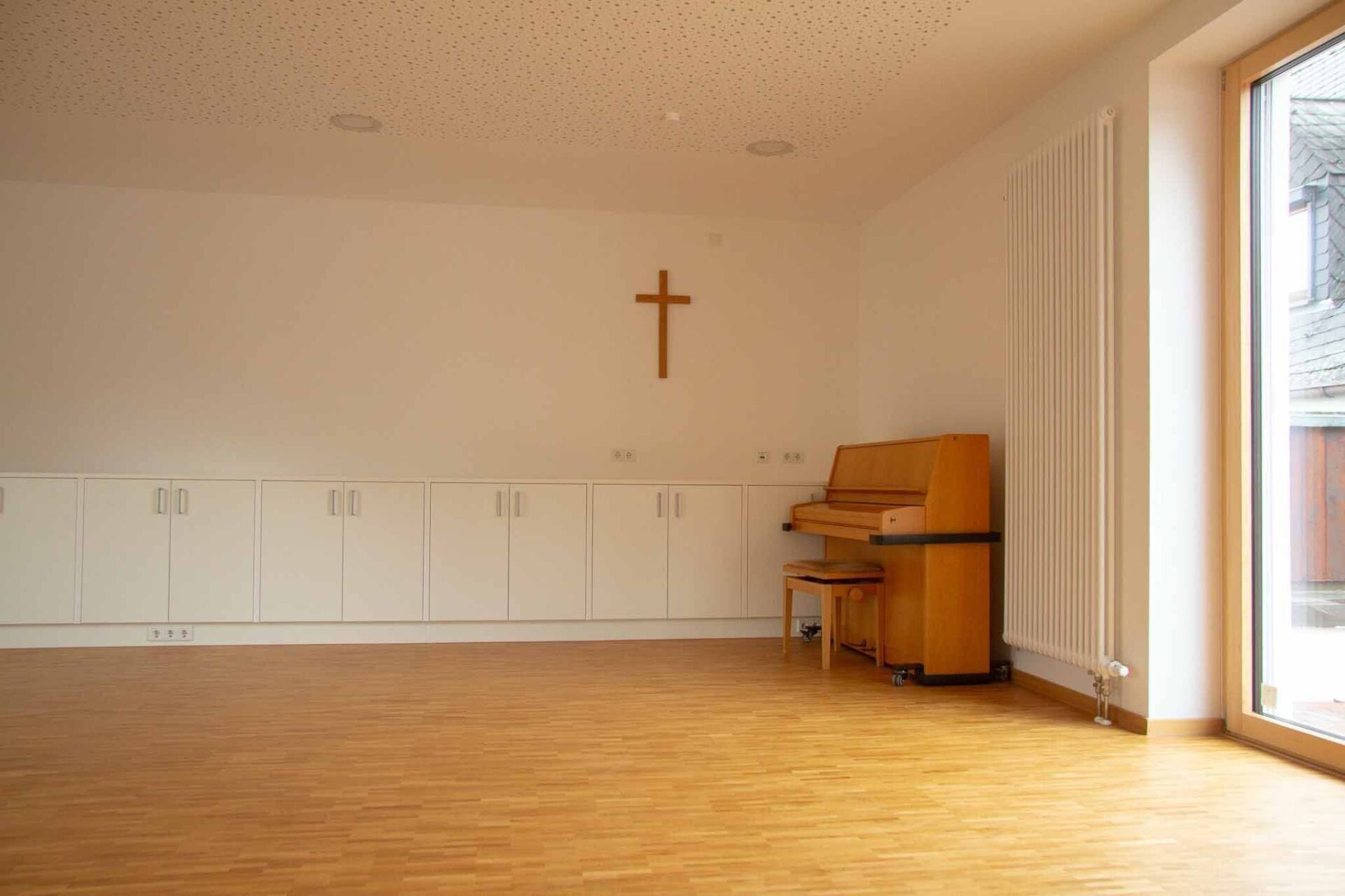 Leerer Raum mit einem Klavier, einem Kreuz an der Wand und einem Fenster.