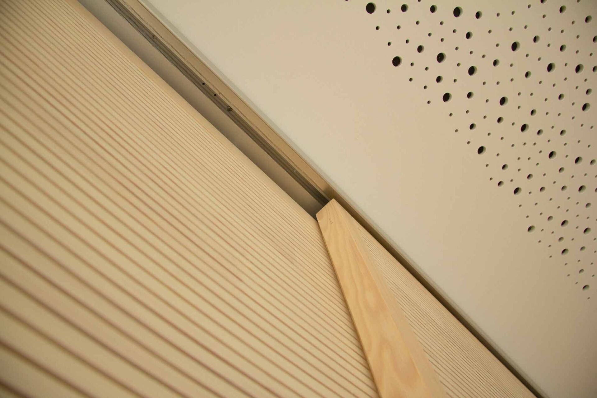 Eine Holzvertäfelung trifft auf eine perforierte weiße Decke, getrennt durch eine Metallschiene.