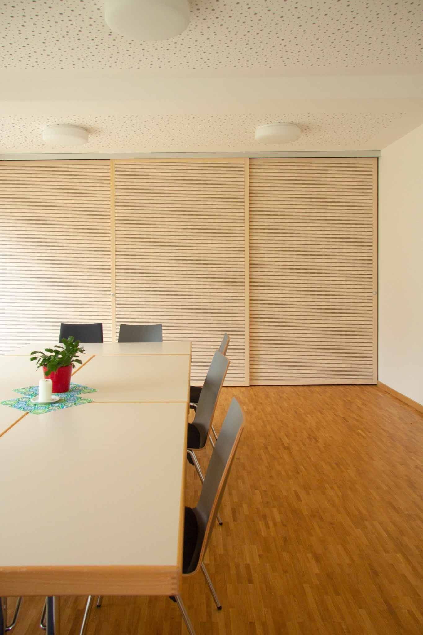 Besprechungsraum mit langem Tisch, Stühlen, Fensterverkleidungen mit Holzlamellen und Holzboden.