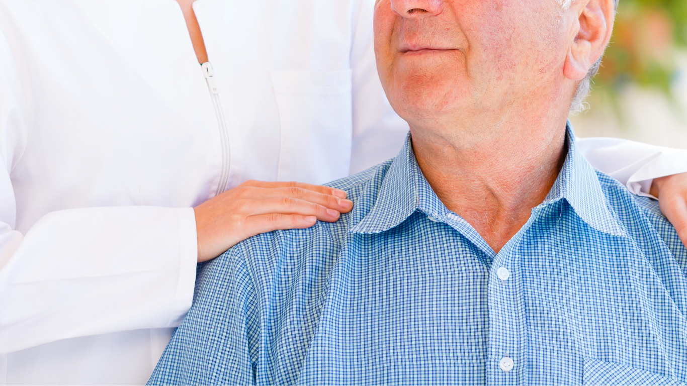 Una persona ayuda a una persona mayor a levantar una tabla marrón, en el interior, cerca de una puerta de madera, mientras la persona mayor viste una camisa naranja.