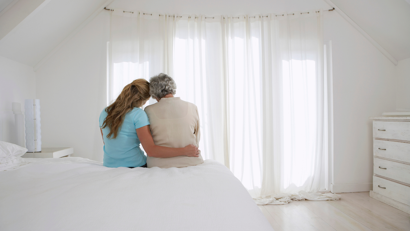 Un trabajador sanitario con uniforme y mascarilla revisa a una persona mayor en un sofá, dentro de una casa.