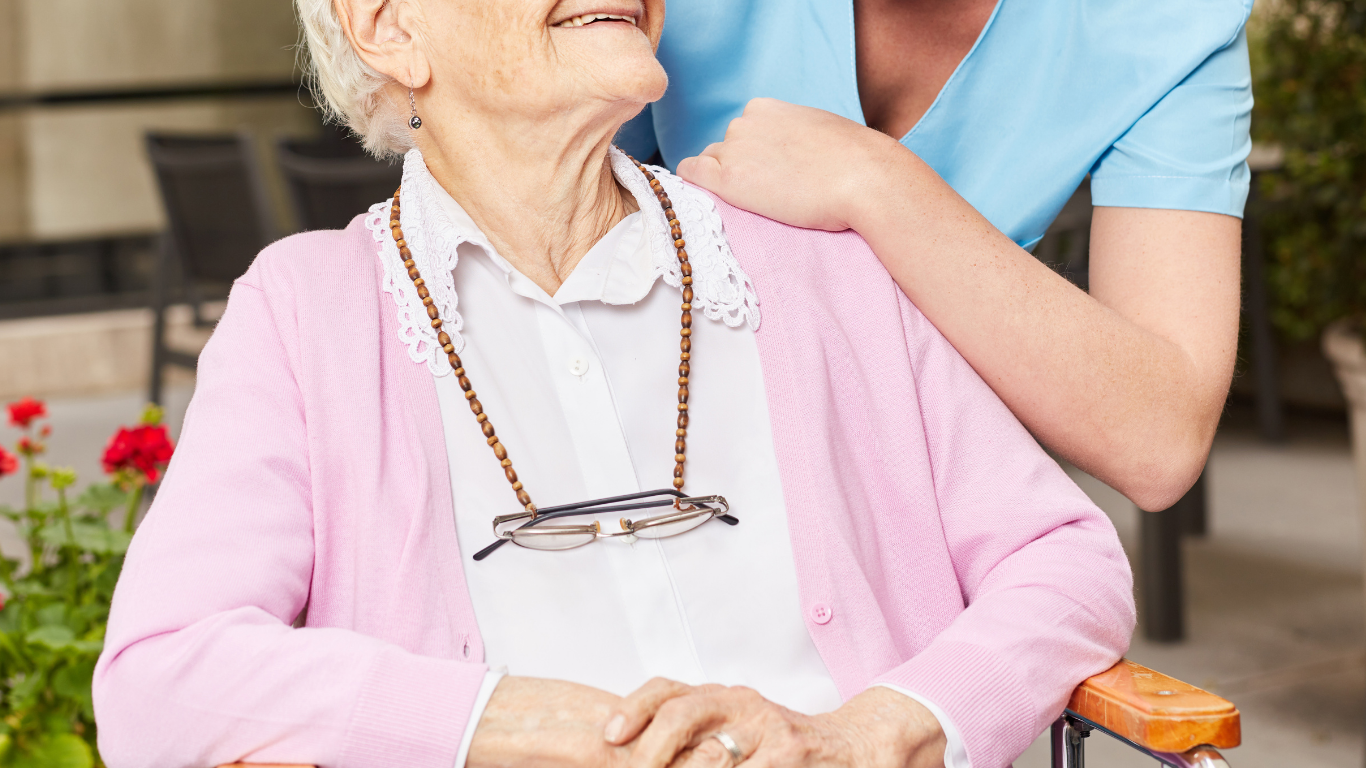 Cuidador ayudando a una persona mayor con un suéter tipo cárdigan en una habitación soleada.