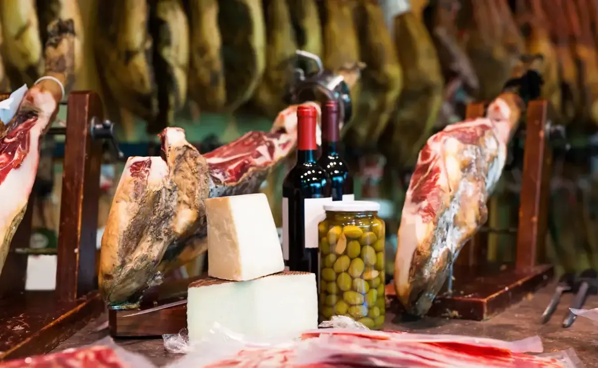 Jamones curados colgados, con jamón en lonchas, queso, aceitunas y botellas de vino tinto sobre un mostrador de madera.