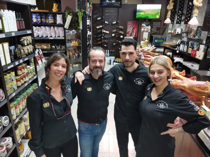 Cuatro personas con camisas negras posan dentro de una tienda con estanterías de productos alimenticios y embutidos.