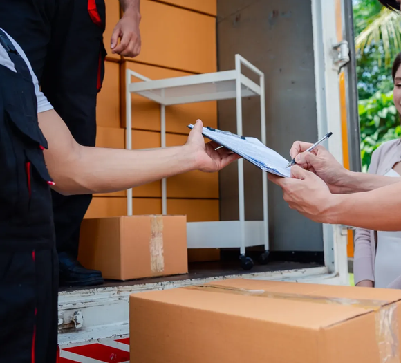 Repartidor entregando un portapapeles a un cliente junto a un camión de mudanzas y cajas de cartón.