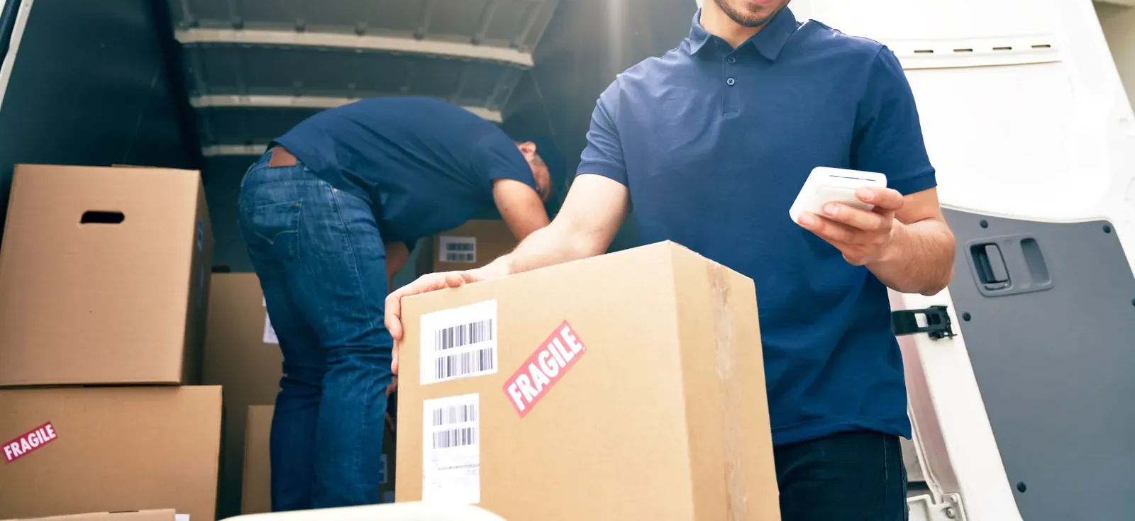 Dos repartidores cargando cajas de cartón en un camión; uno de ellos sostiene un paquete con un escáner.