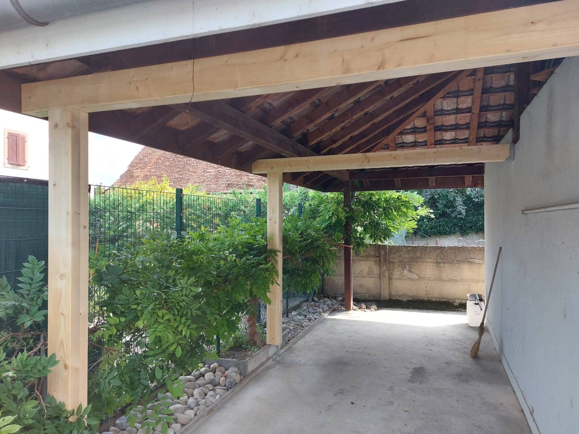 Abri voiture avec poutres en bois et toit de tuiles, faisant extension d'un bâtiment.