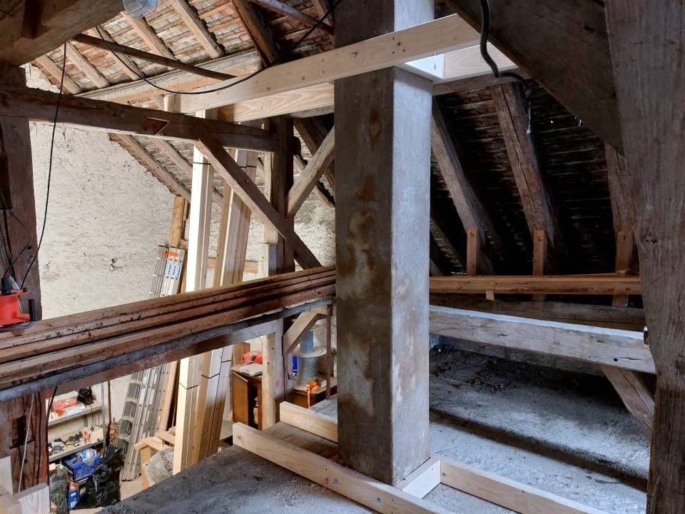 Vue intérieure d'un grenier avec poutres en bois apparentes, une colonne centrale en béton et des matériaux de construction.