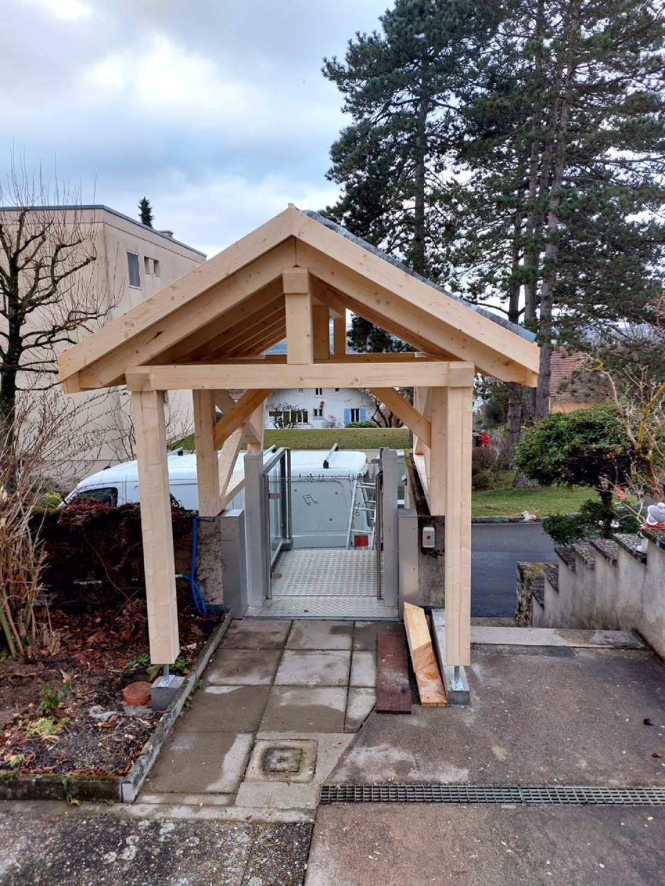 Entrée d'un abri de voiture en bois avec allée pavée, portail et cadre en bois clair, arbres en arrière-plan.