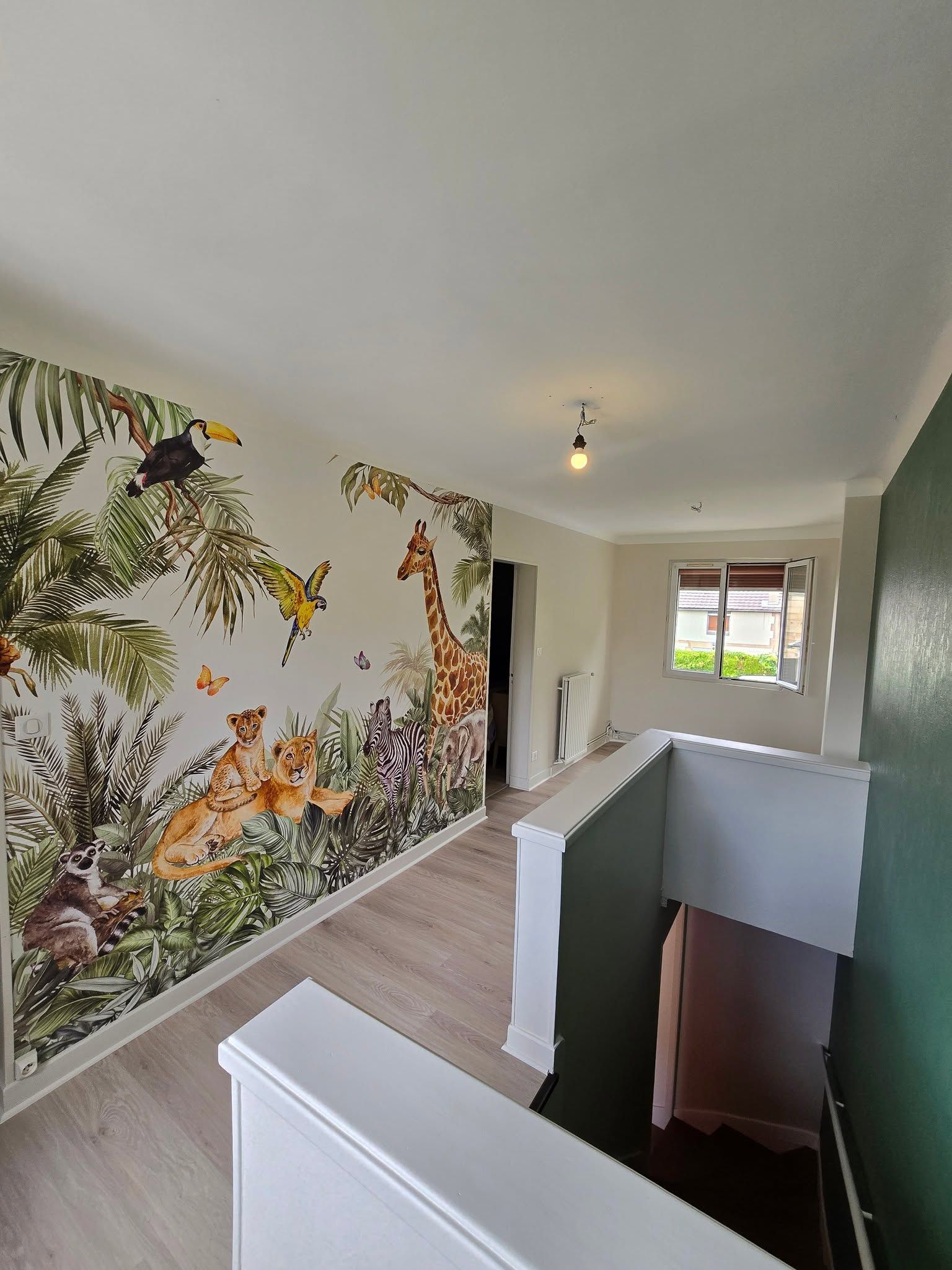 Couloir lumineux avec une fresque tropicale, un parquet clair et une rambarde donnant sur l’escalier, avec une fenêtre au fond