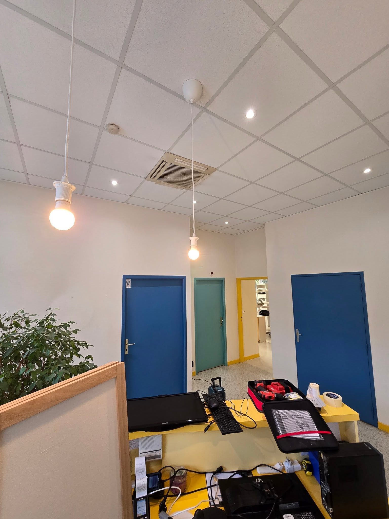 Couloir de bureau avec des portes bleues, des luminaires suspendus et un petit coin salon avec des sacs et des papiers.