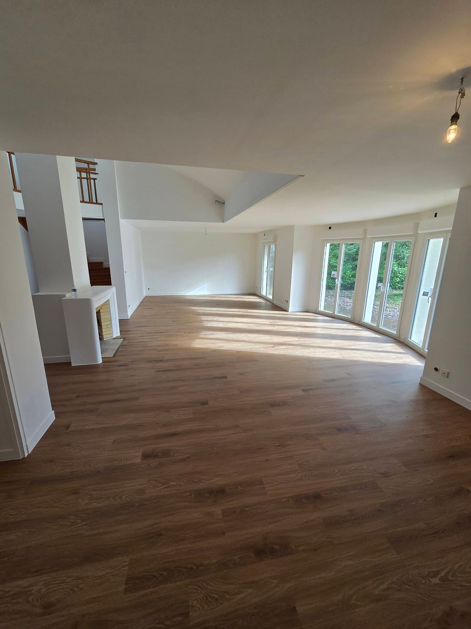 Salon moderne vide avec parquet, murs blancs et grandes fenêtres laissant entrer la lumière du soleil.